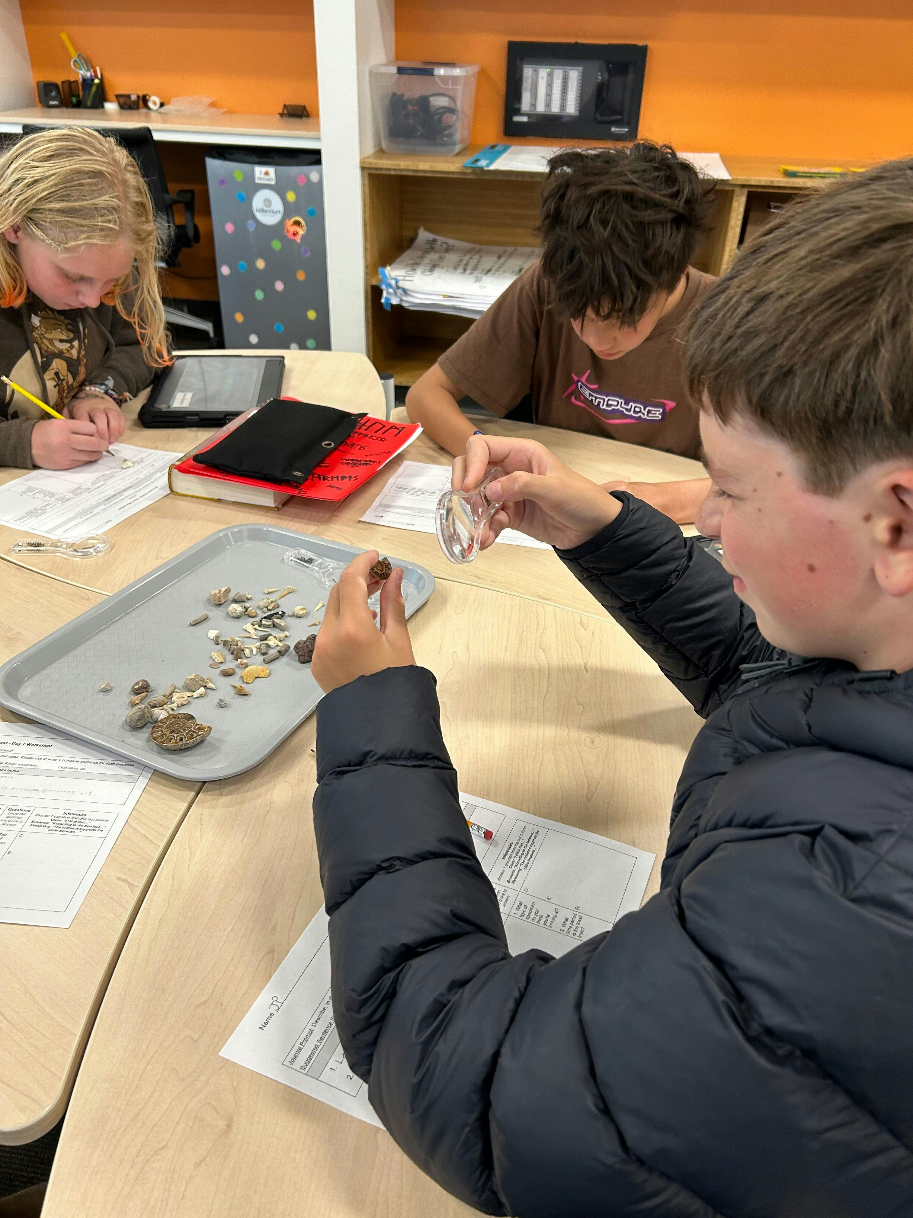 Three children in a classroom examining small rocks and fossils on a tray