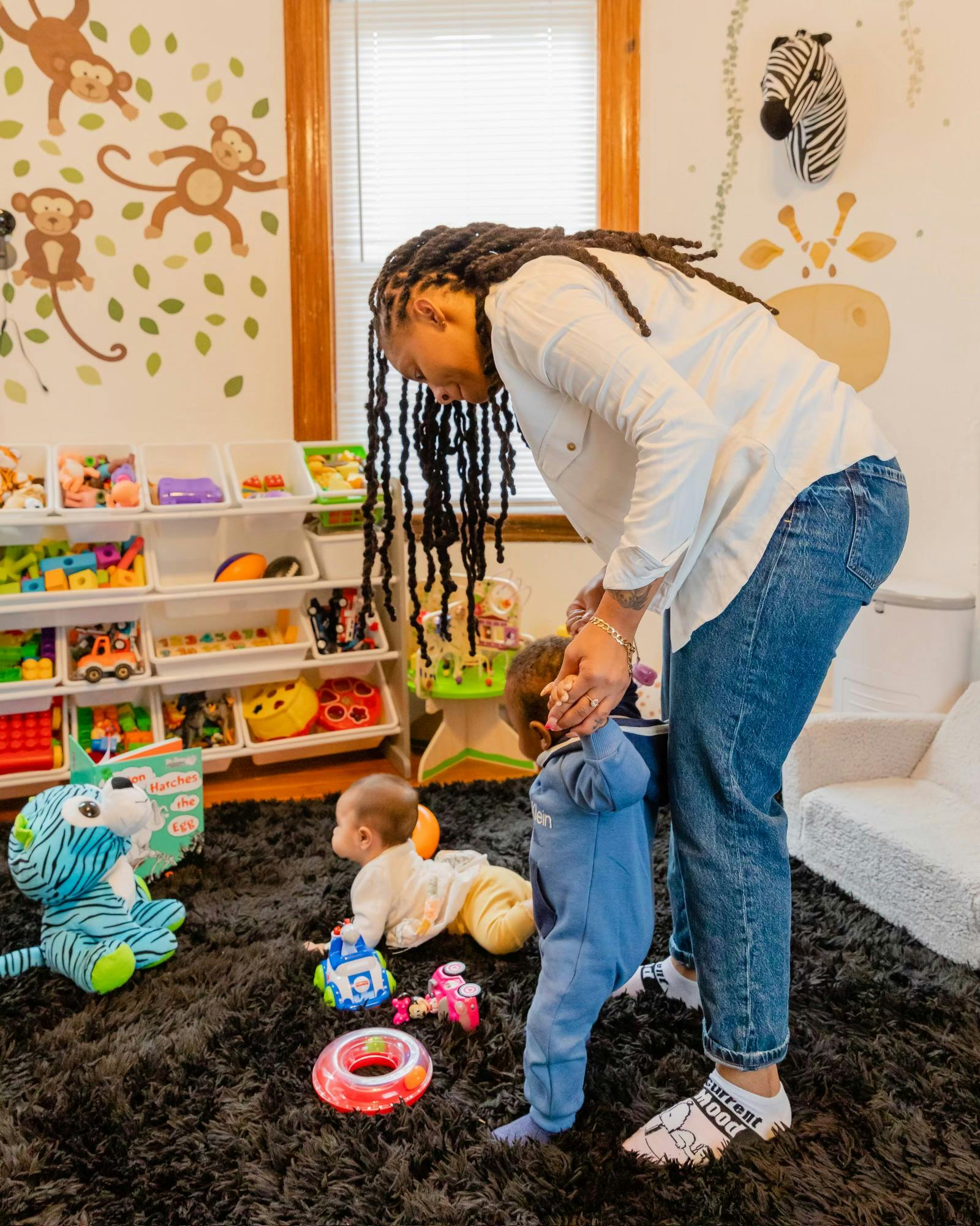 Person helping a child stand while another child crawls on the floor
