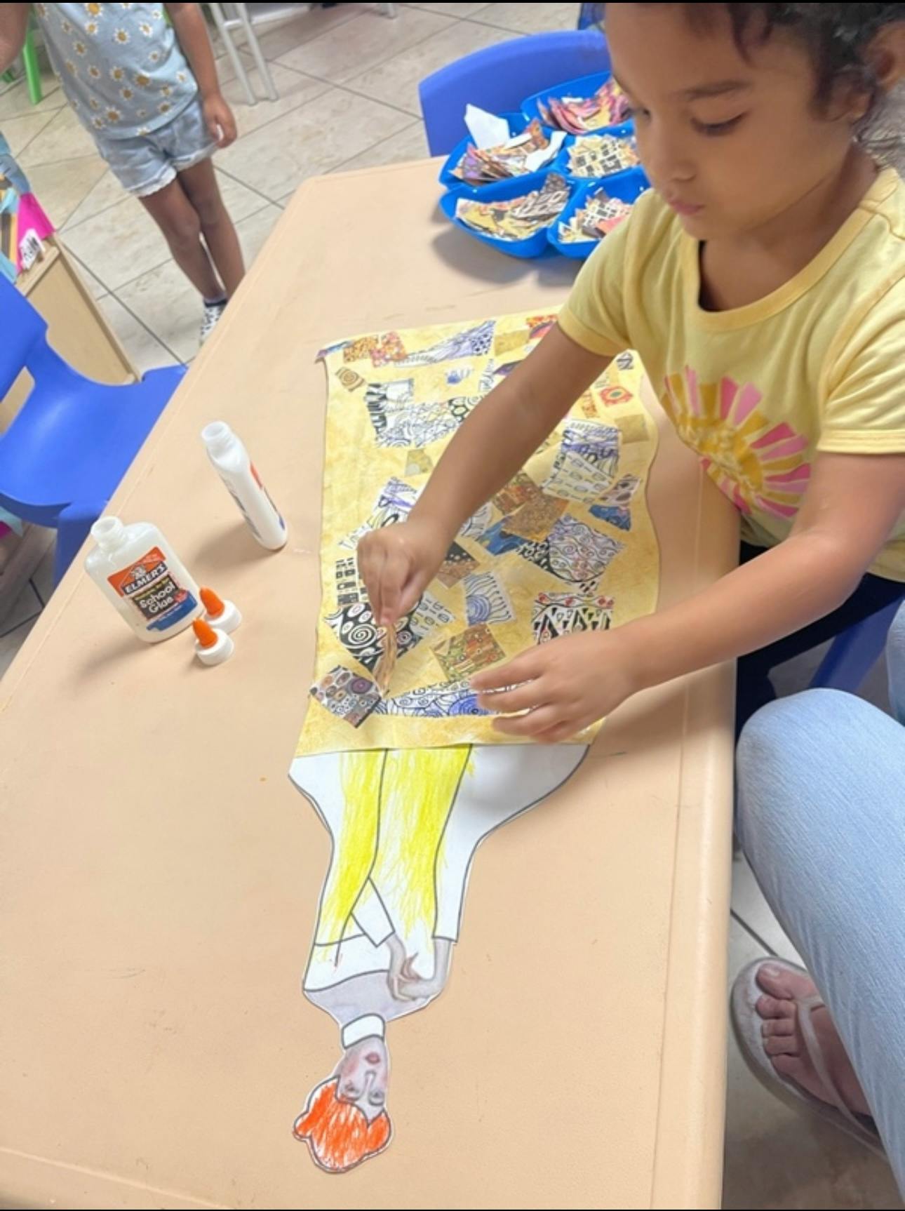 Child gluing paper collage pieces onto a cutout illustration at a table