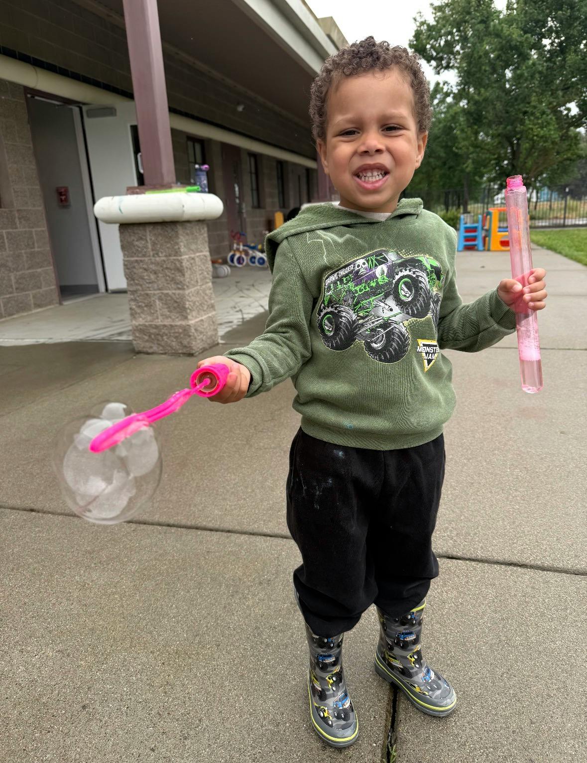 Child in a green sweatshirt and boots holding a bubble wand and bottle