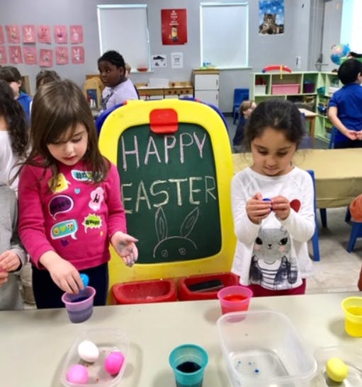 Two children decorating eggs in a classroom with a "Happy Easter" sign