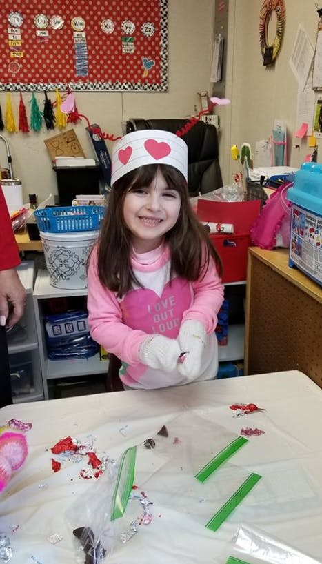 Child in a paper hat with hearts, wearing gloves, standing at a table