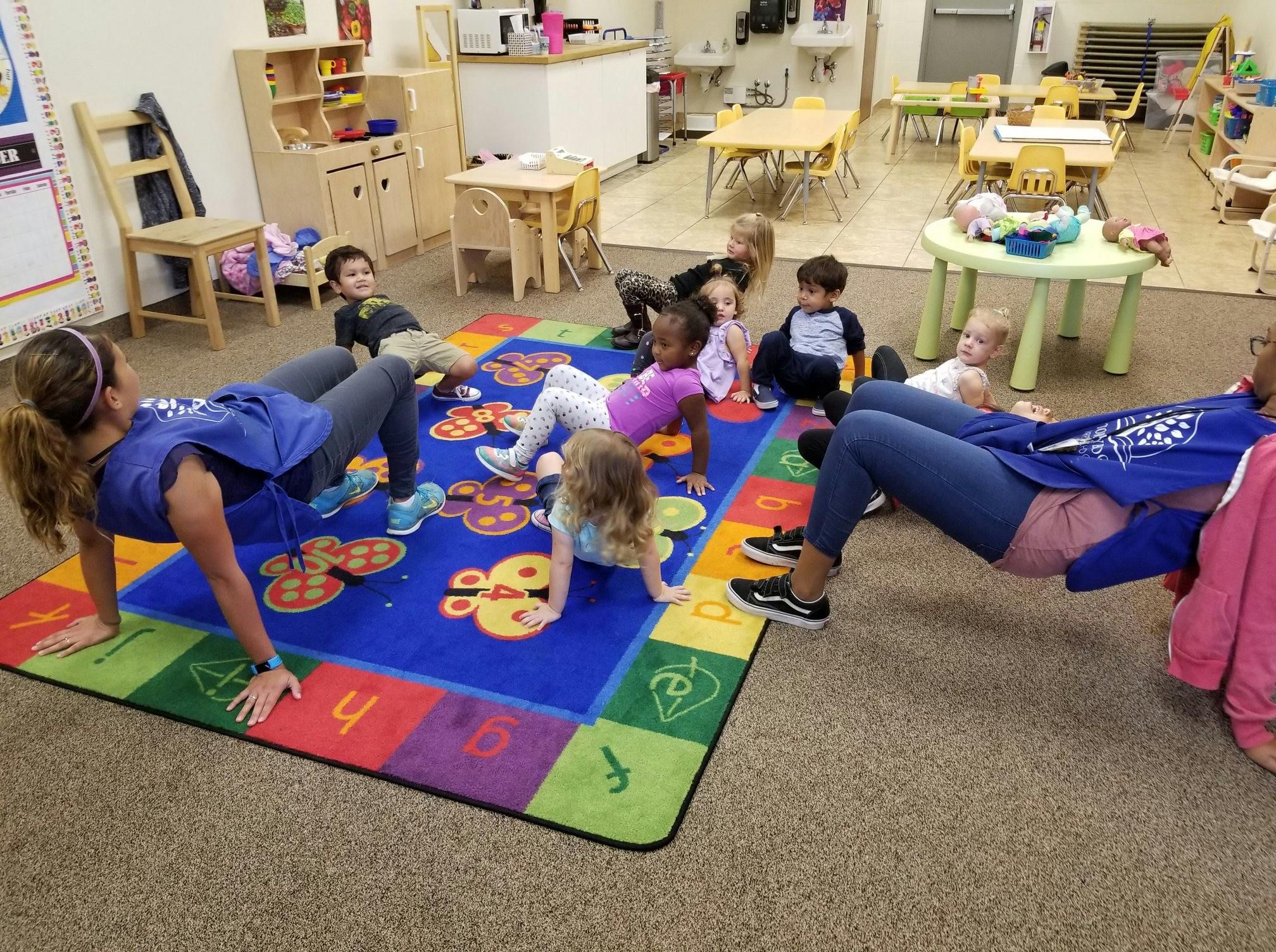 Two people and multiple children doing exercises on a colorful rug in a classroom