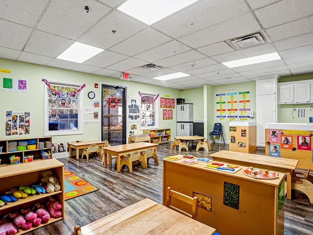 An empty classroom with small wooden tables and chairs, toys on shelves