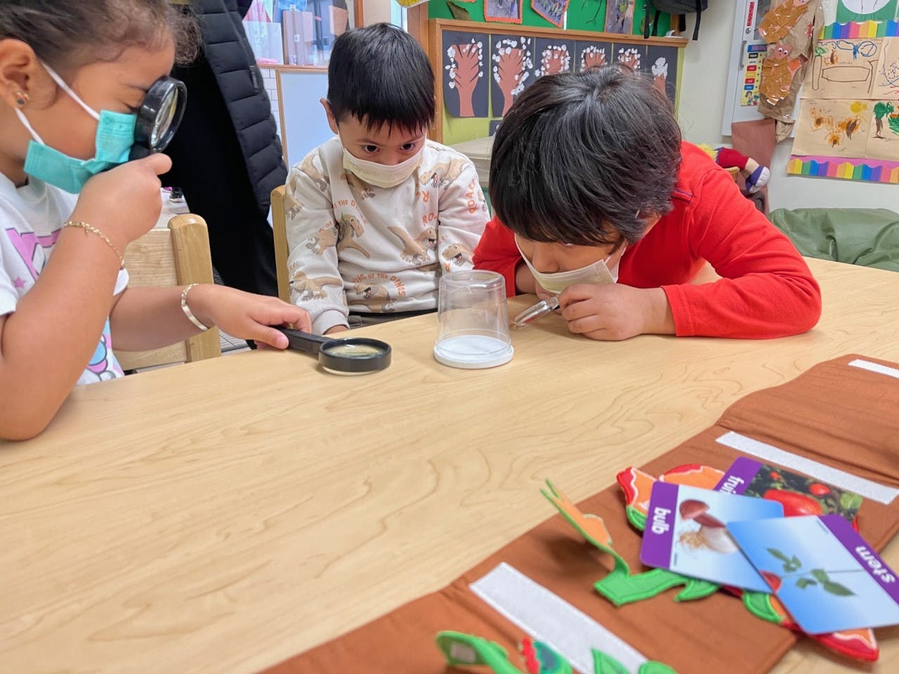 Three children wearing masks looking at objects with magnifying glasses at a table