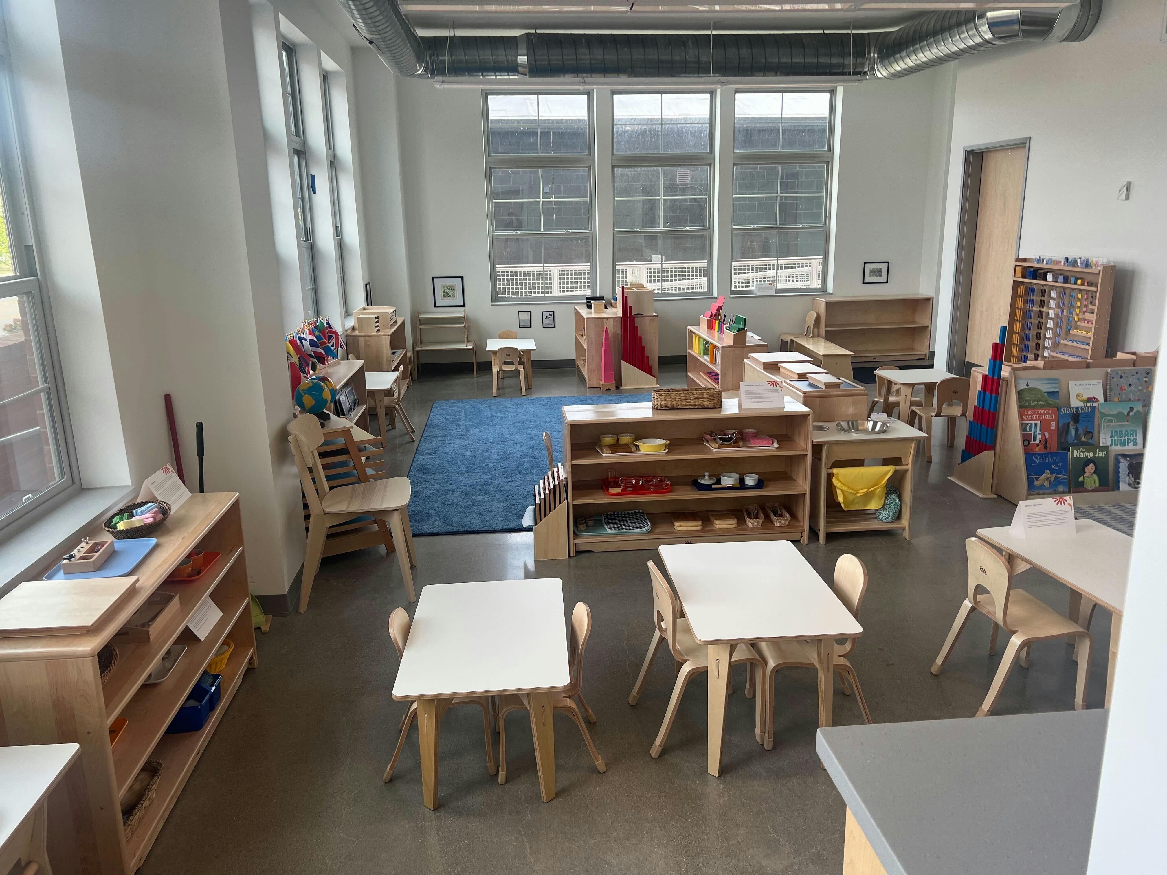 Classroom with small tables and chairs, shelves with learning materials, and a blue rug