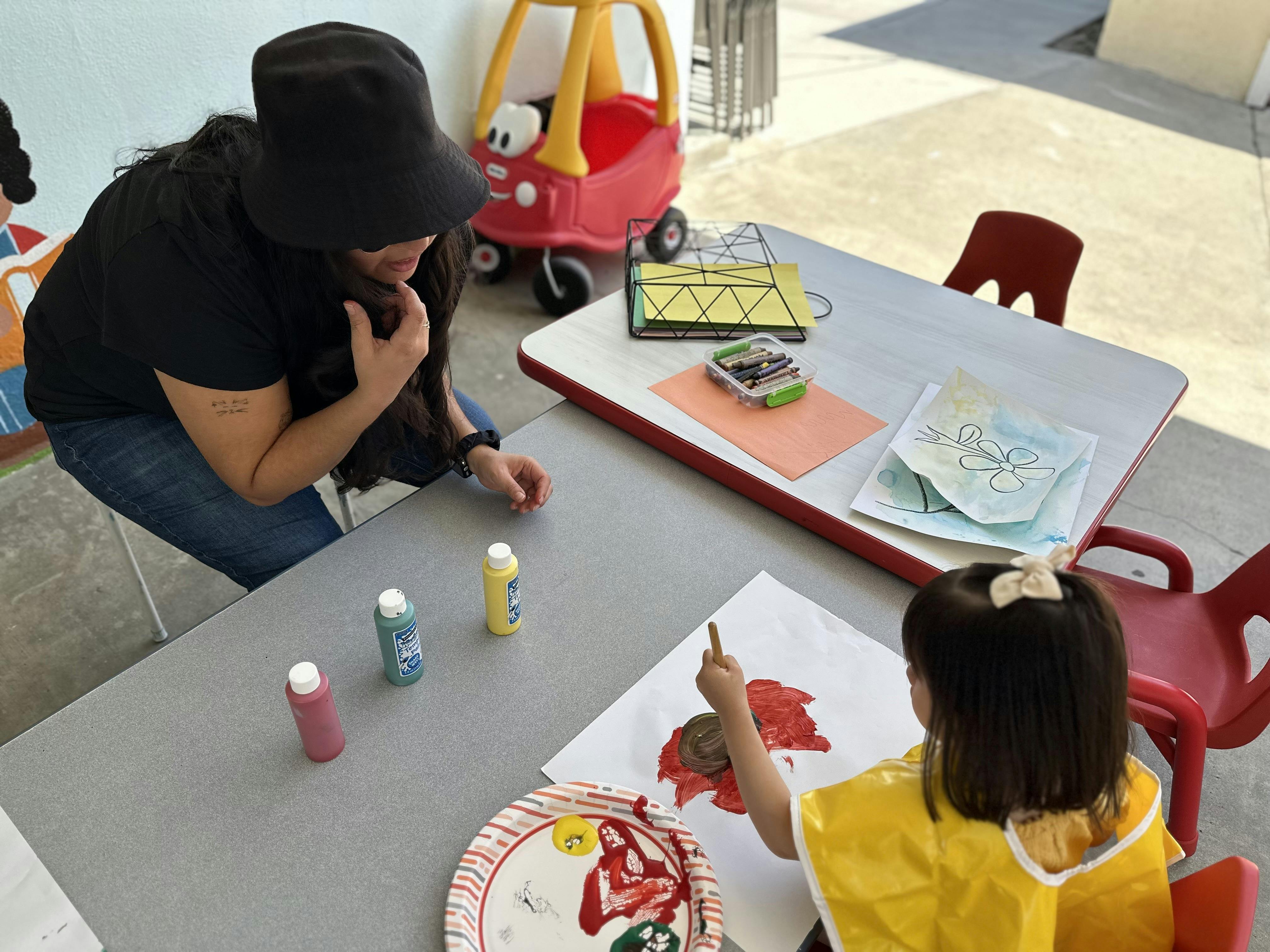 Person in a hat watching a child paint at a table