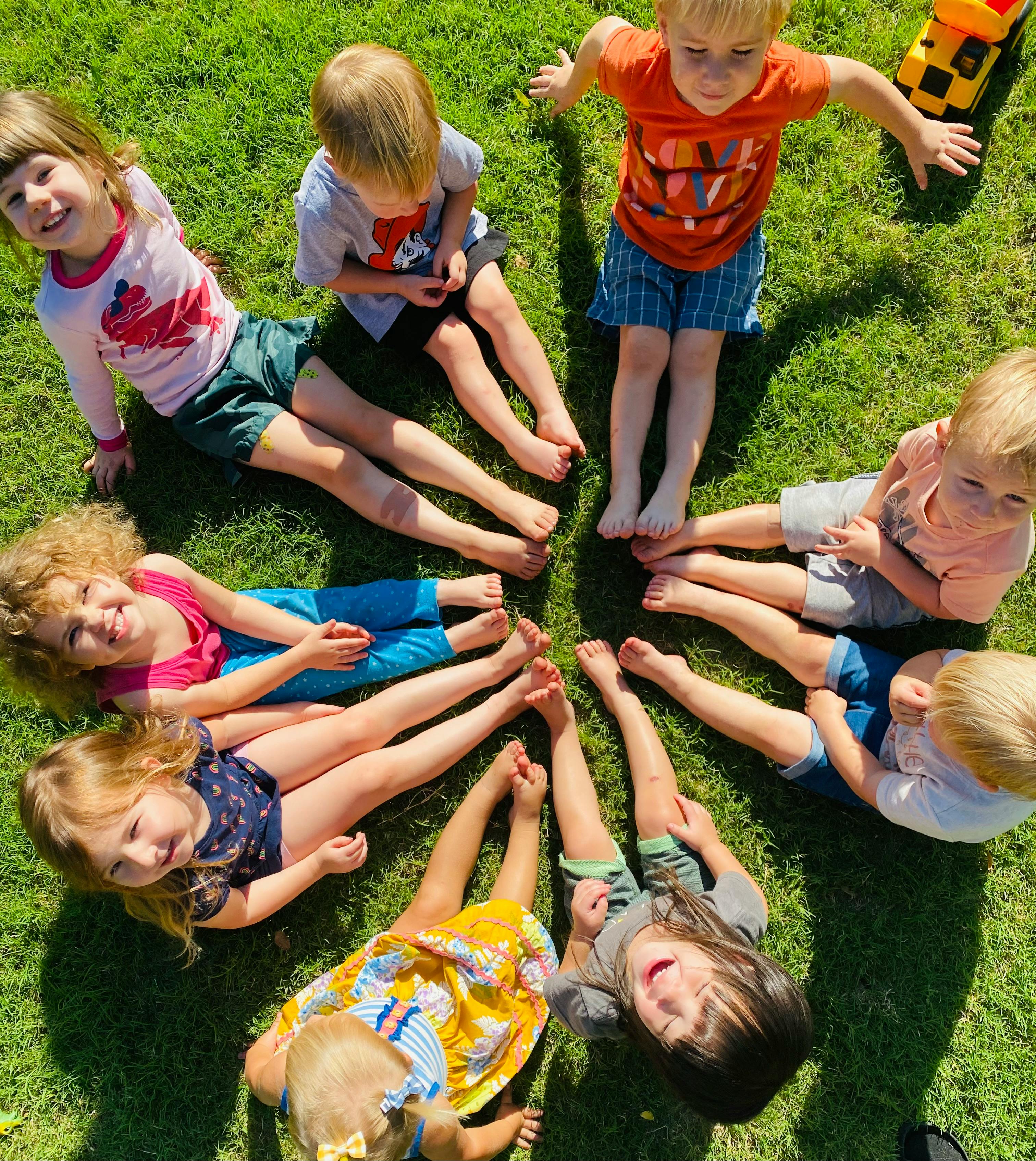 Children sitting in a circle on grass with their feet touching in the middle