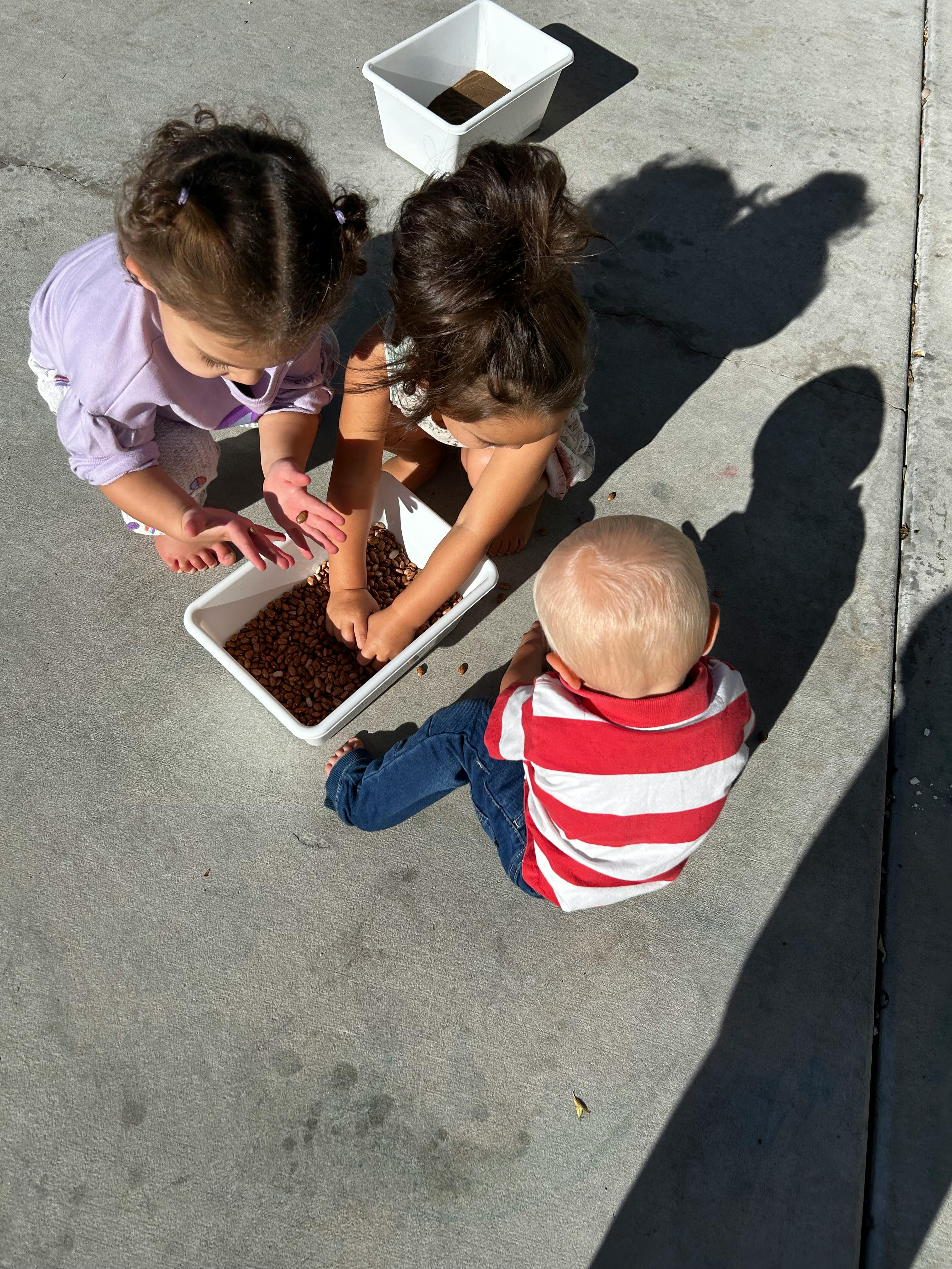 Two children playing with brown material in a white bin, rectangular container