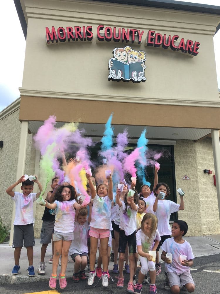Children jumping and throwing colorful powder in front of a building with "MORRIS COUNTY EDUCARE" sign