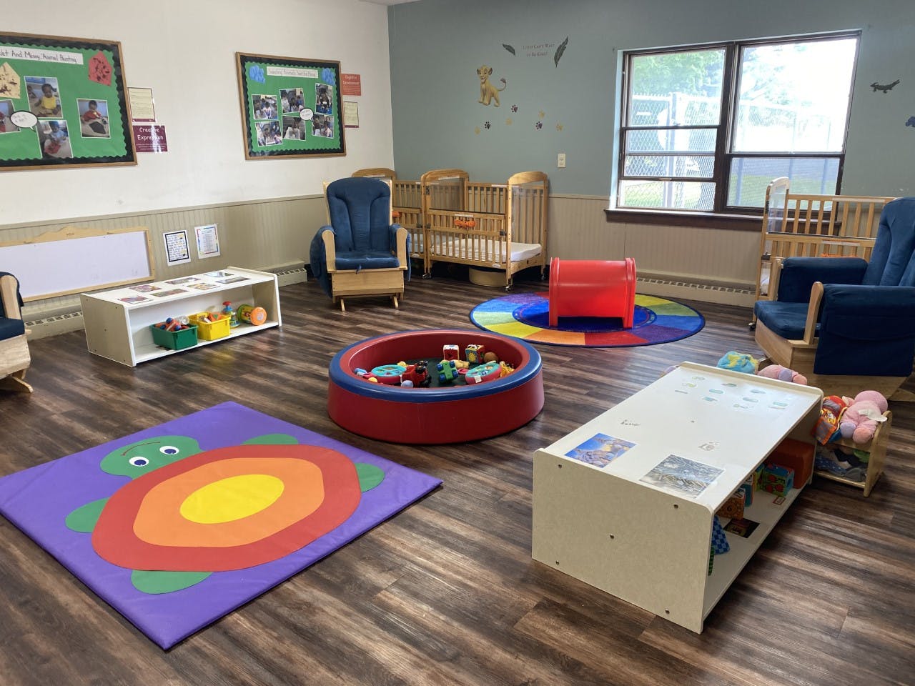 A colorful childcare room with cribs, rocking chairs, play mats, and toys
