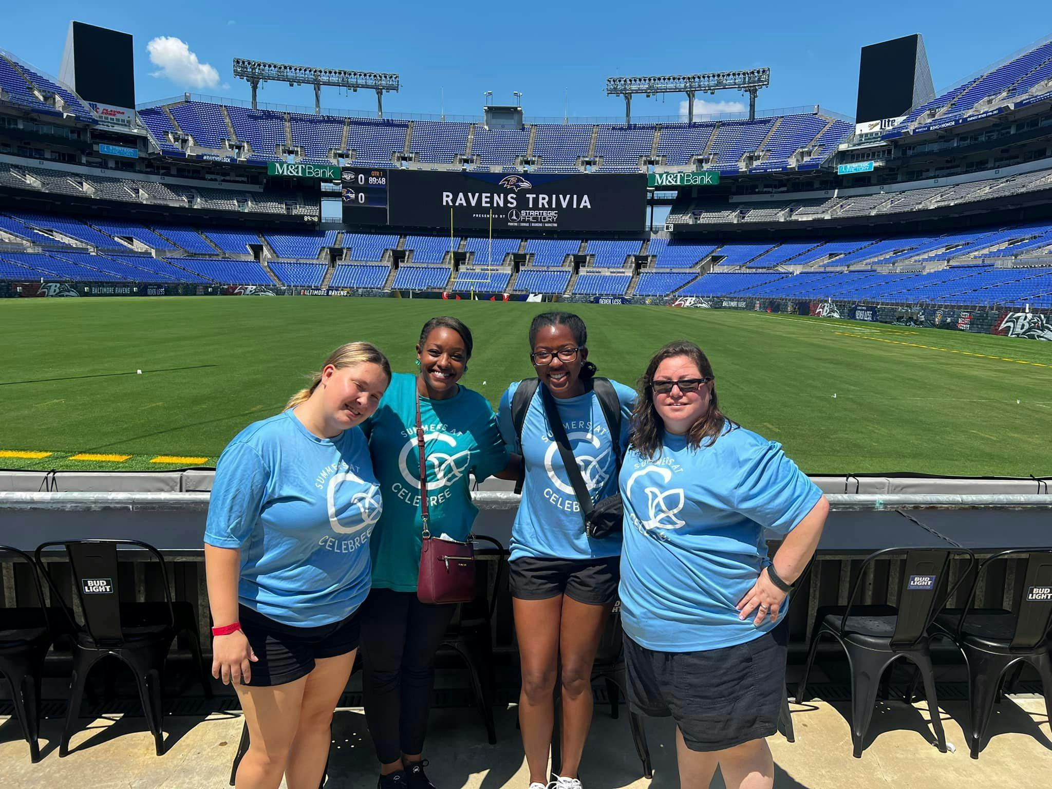 Four people standing in front of a stadium field with "RAVENS TRIVIA" on a screen