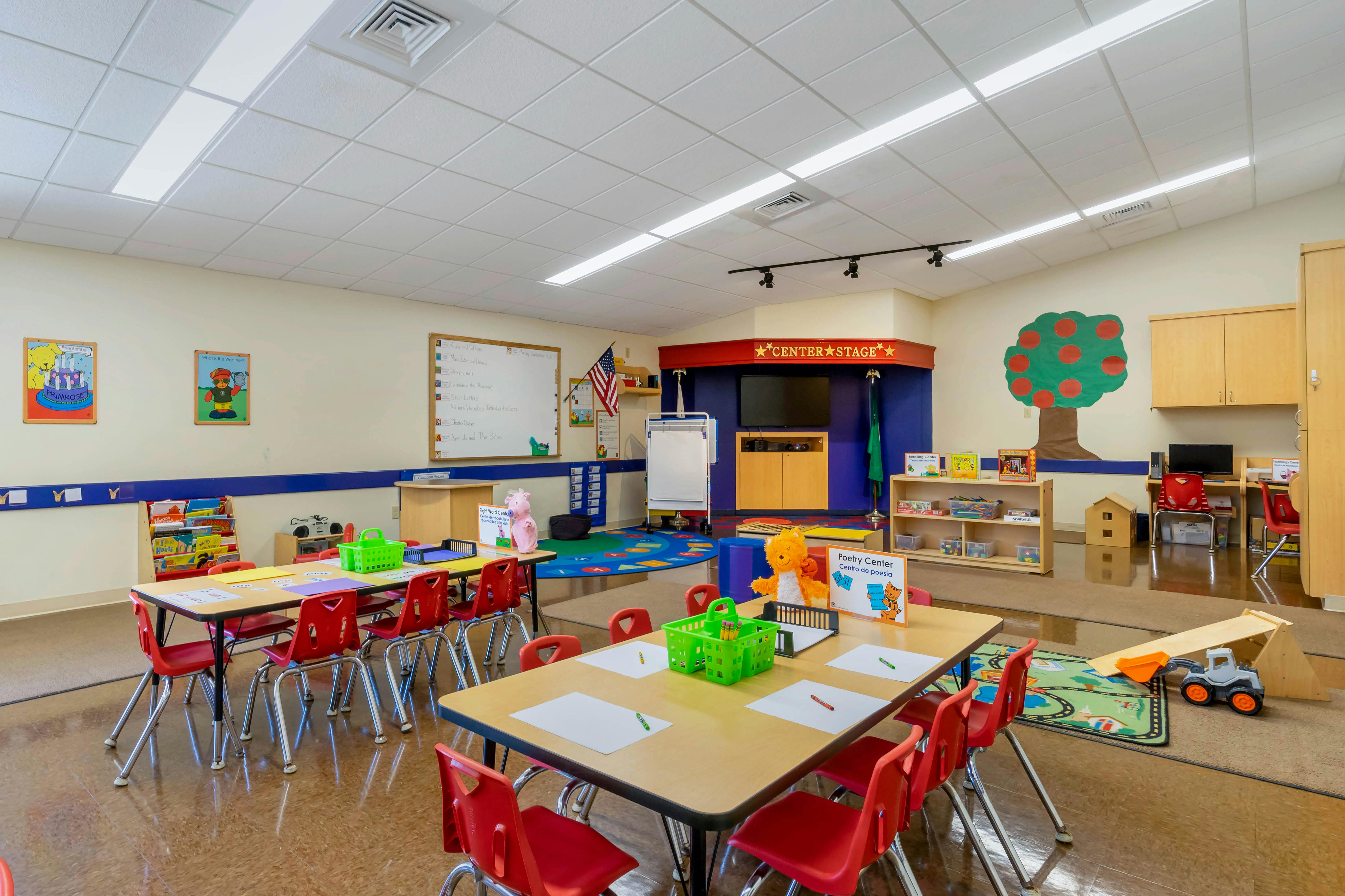 Classroom with small tables and red chairs, a stage area, and activity centers