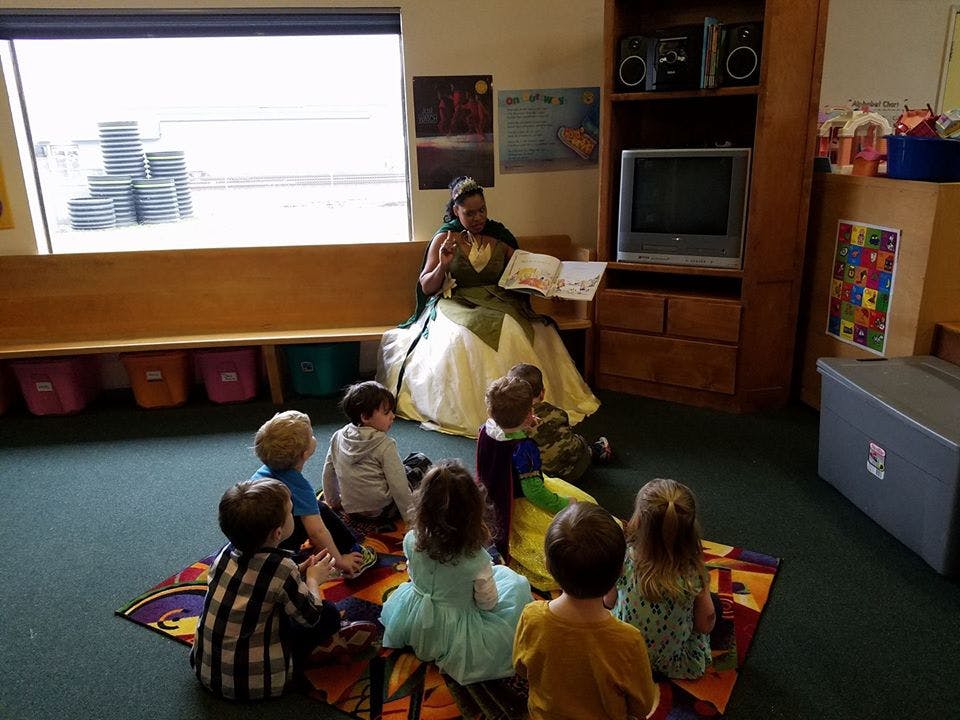 Person in a dress reading a book to a group of children sitting on a rug