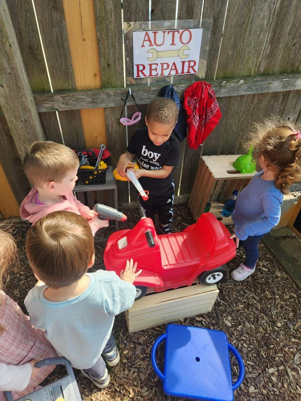 Children playing with toy tools around a red toy car, "AUTO REPAIR" sign