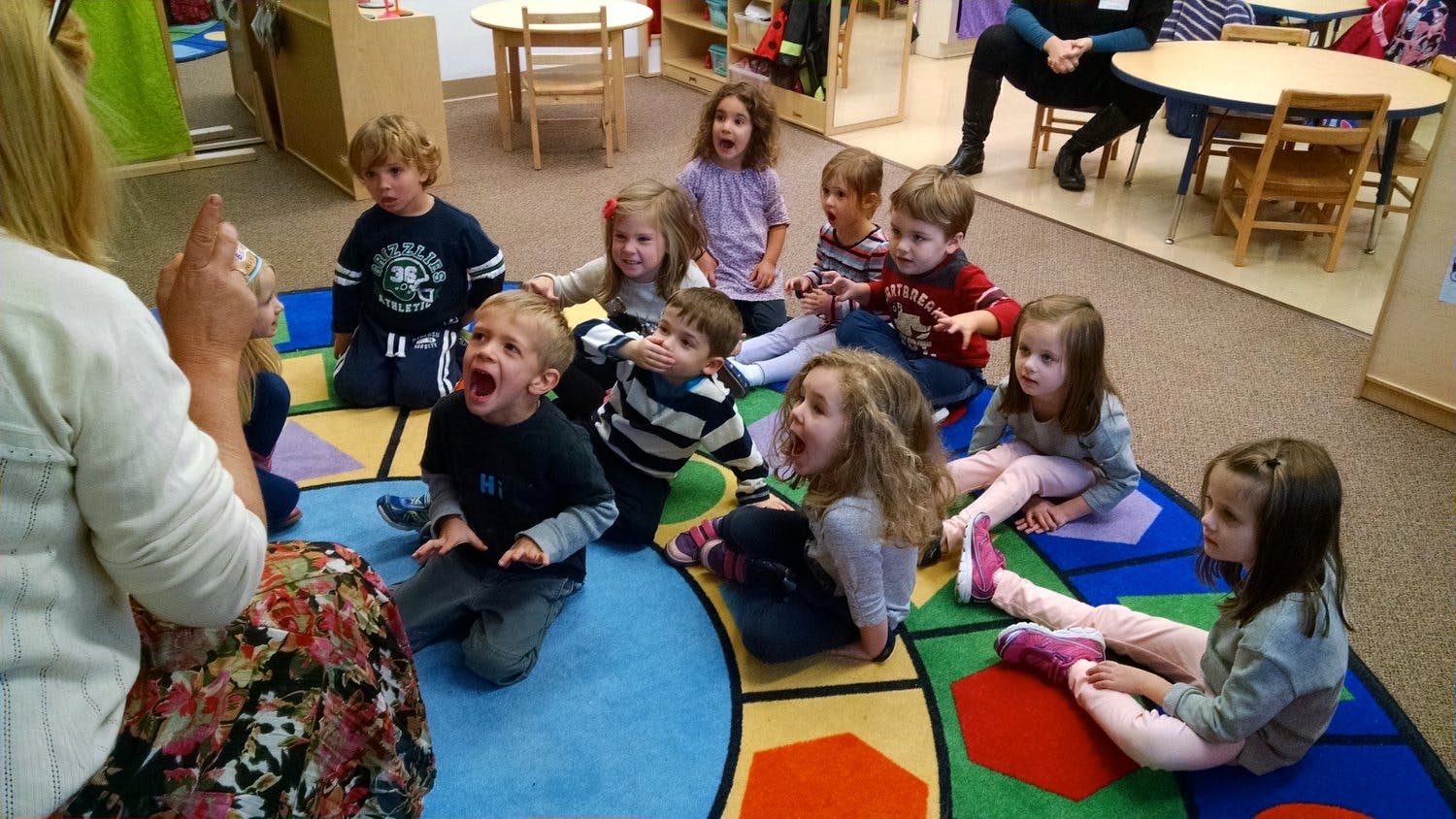 Person leading a group of children sitting on a colorful rug in a classroom