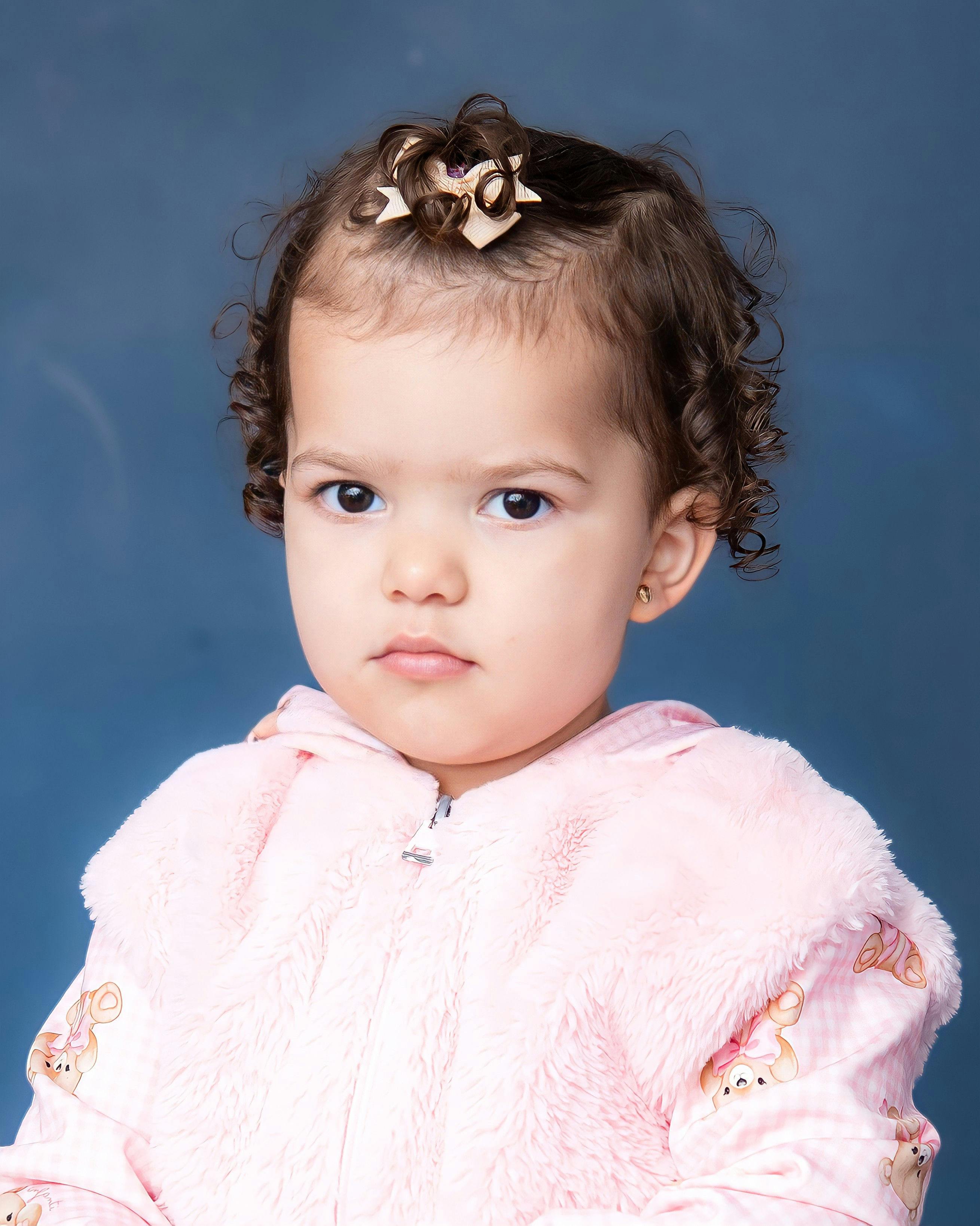Child with curly hair and a bow wearing a pink fuzzy jacket