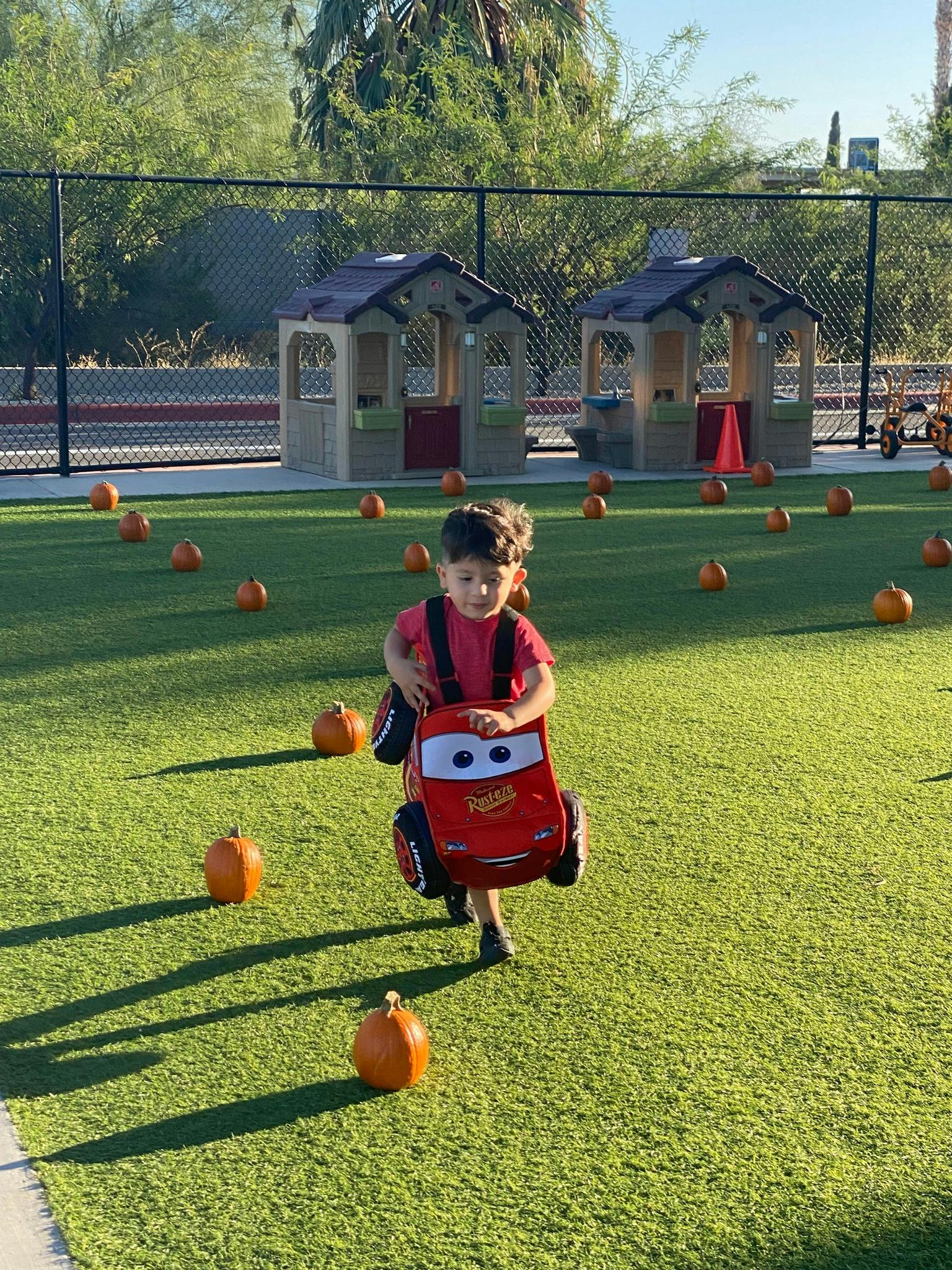 Child in a red shirt and a Lightning McQueen backpack walking on grass with pumpkins