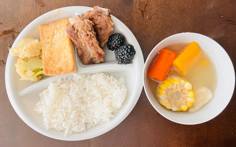 A segmented plate with rice, meat, tofu, cauliflower, and berries next to a bowl of soup
