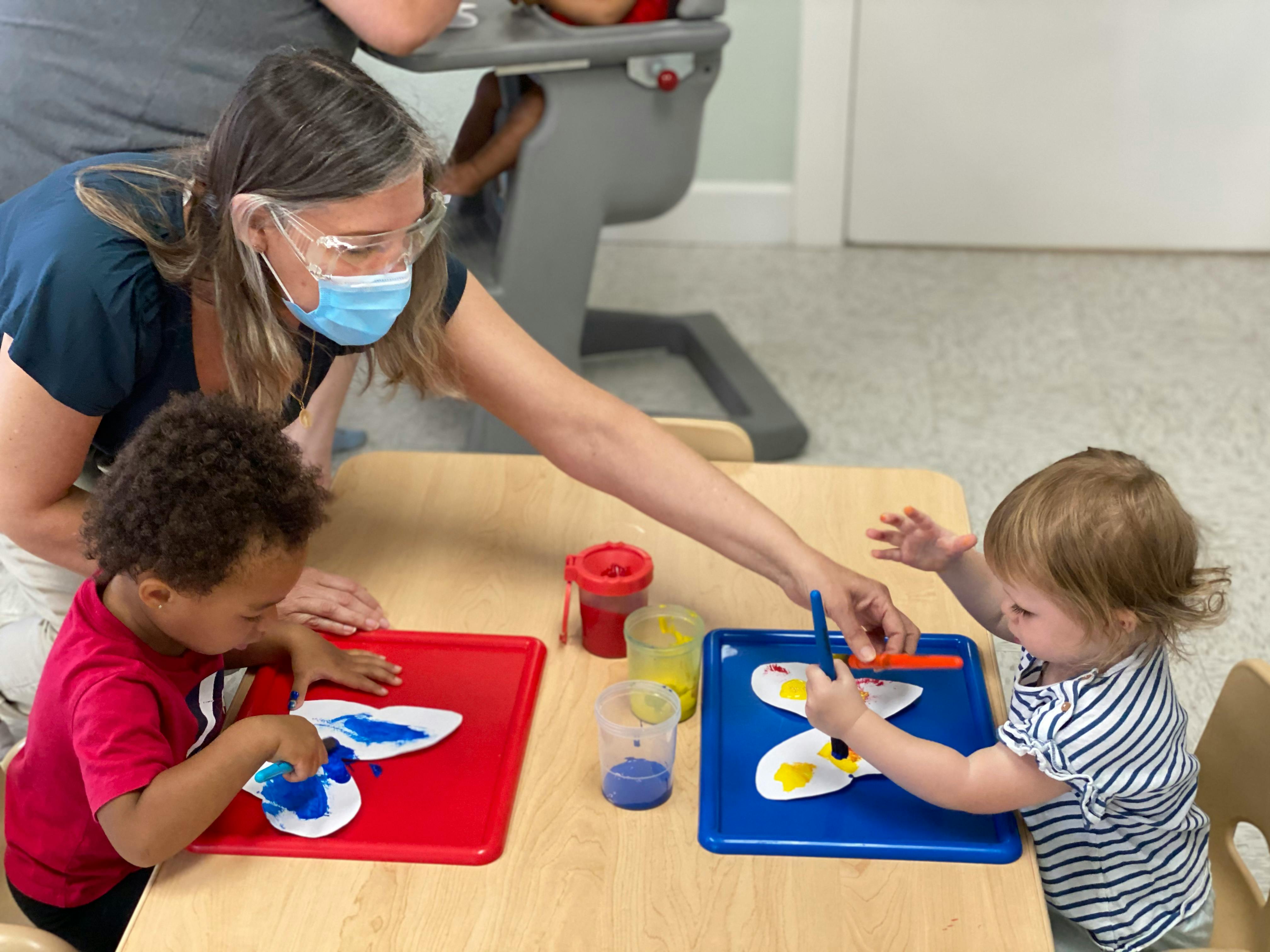 Person in mask and safety glasses helping two children with art projects