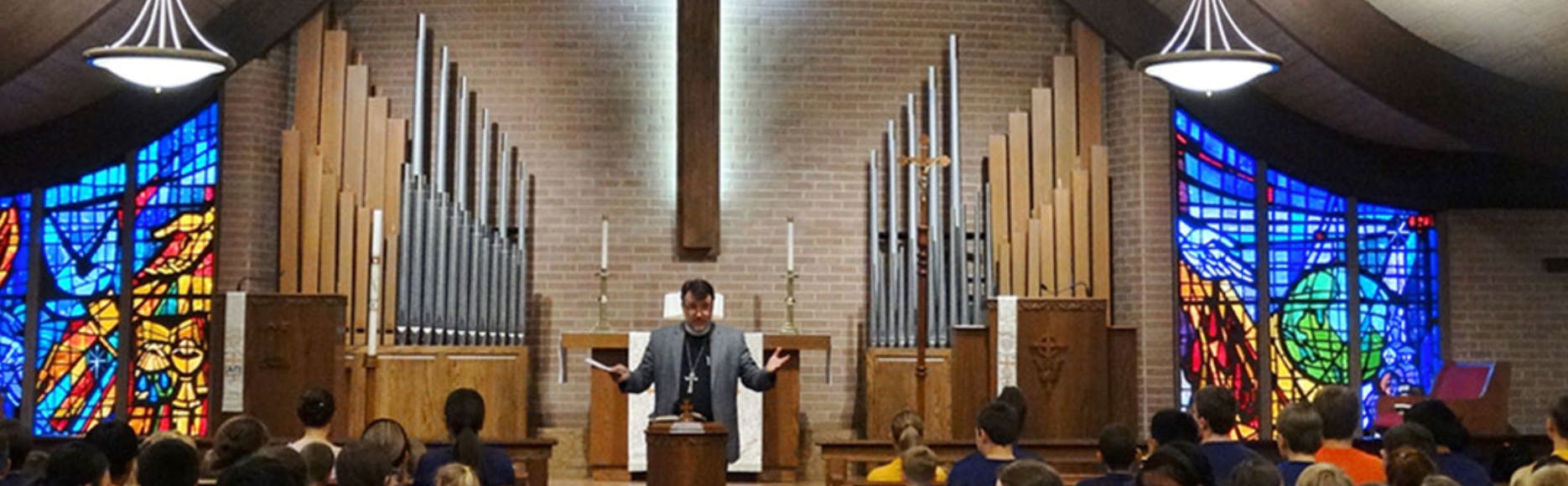 Person speaking at a podium in a church with stained glass windows and organ pipes