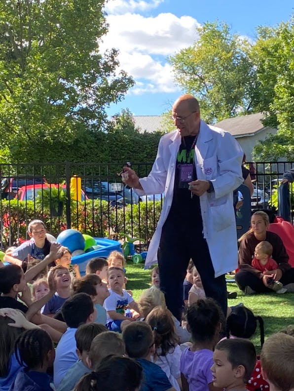 Person in a white lab coat presenting to a group of children outdoors