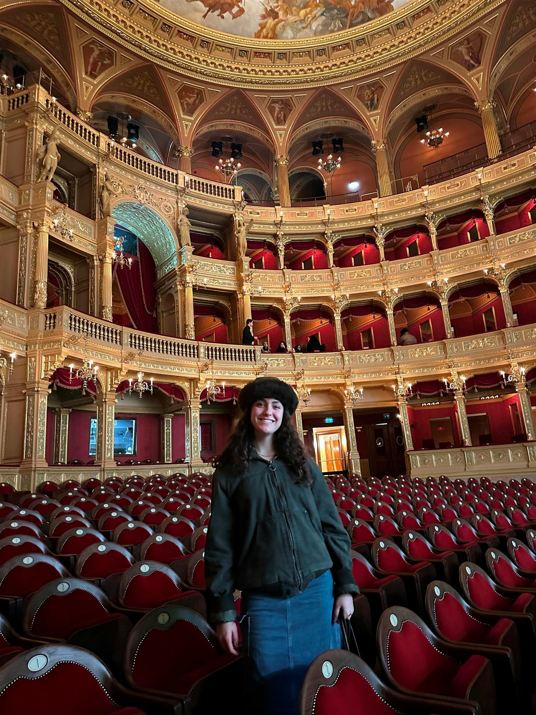 Person with long hair and a hat smiling in an ornate theater with red seats and balconies