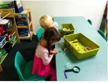 Two children at a table with green bins and a magnifying glass