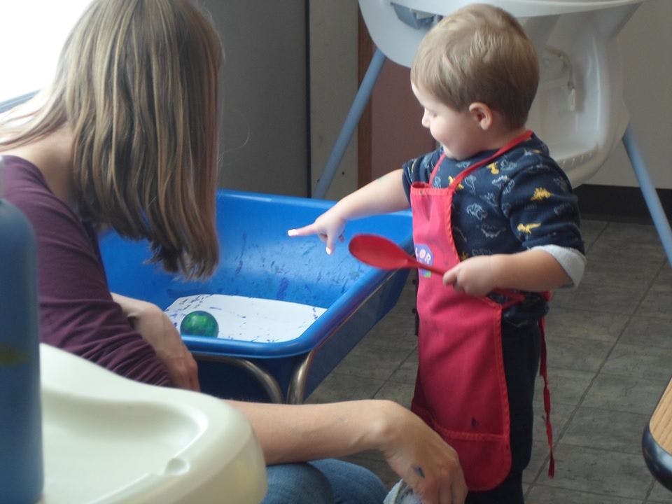 Child in red apron pointing at blue bin with person