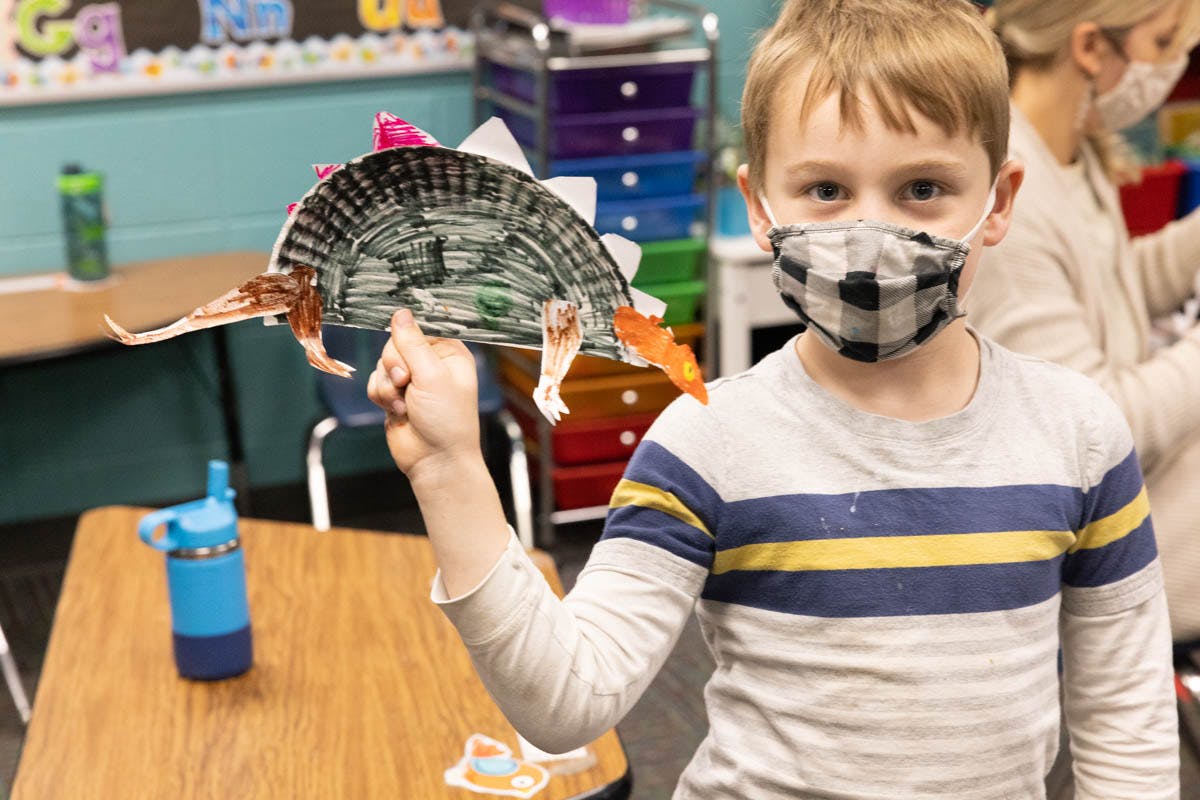 Child in a mask holding up a paper craft in a classroom