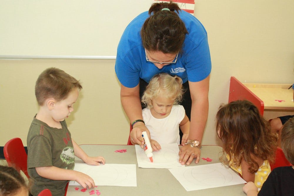 Adult helping a child draw on paper with a marker, other children nearby