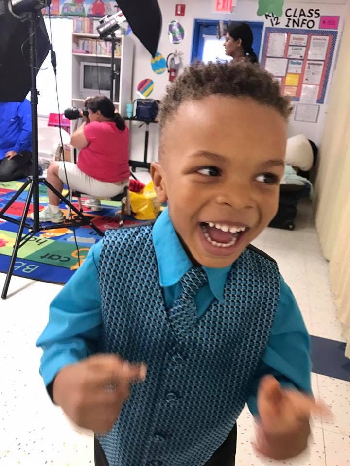 Child in a blue shirt and vest smiling, person filming in background