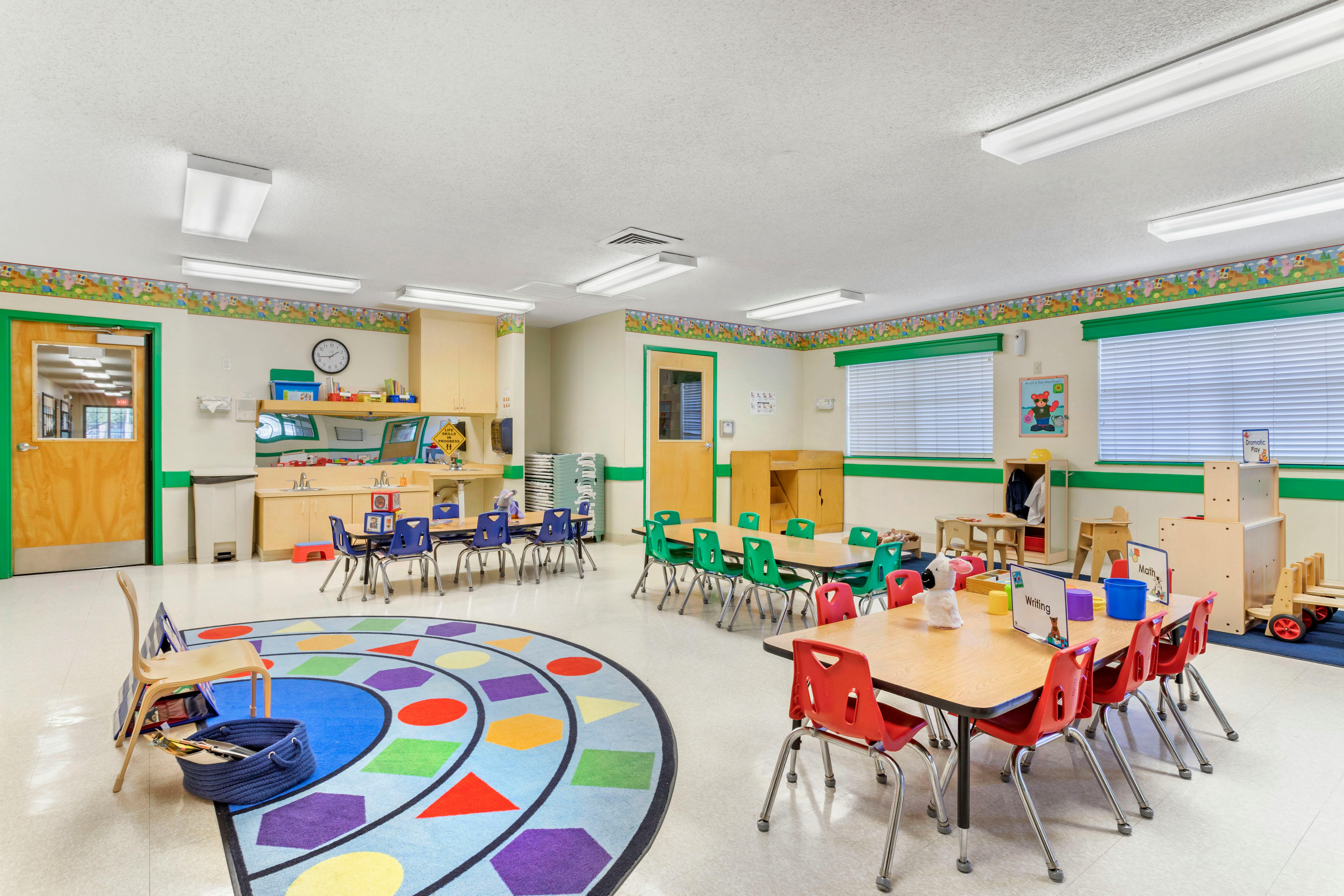 Colorful classroom with child-sized tables and chairs, a sink counter, and a shape rug