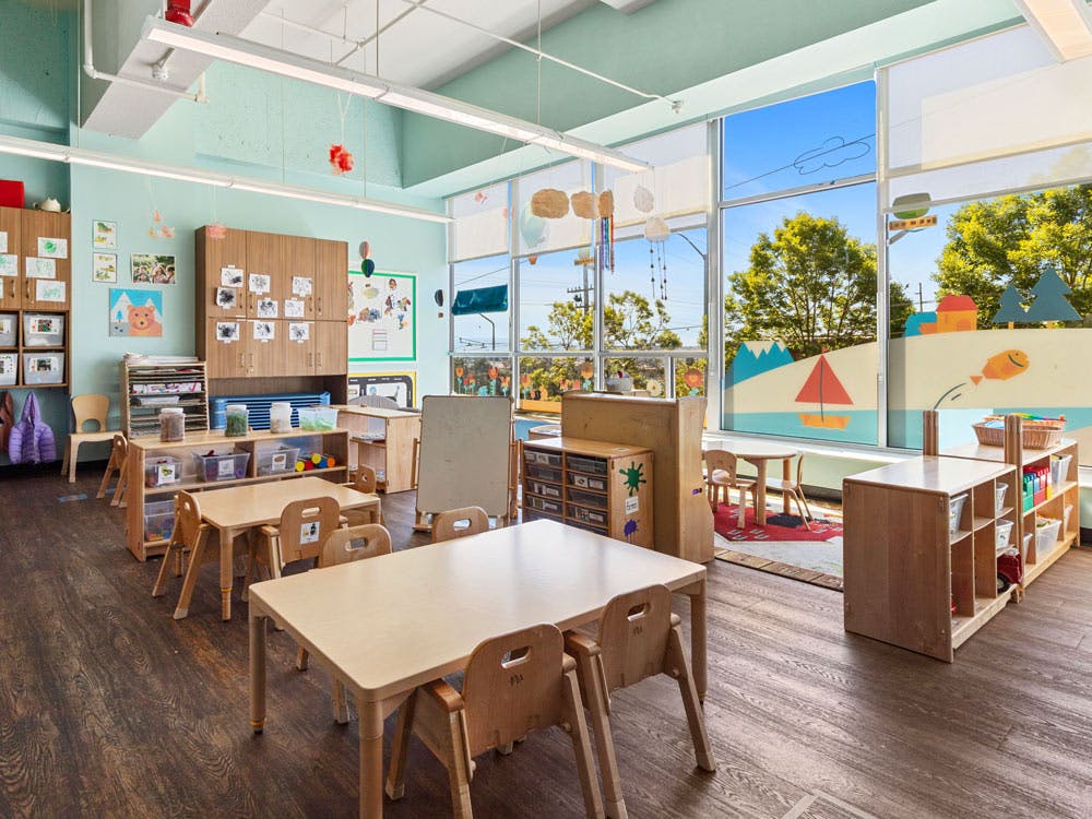 Classroom with small wooden tables and chairs, large windows with decorated panels