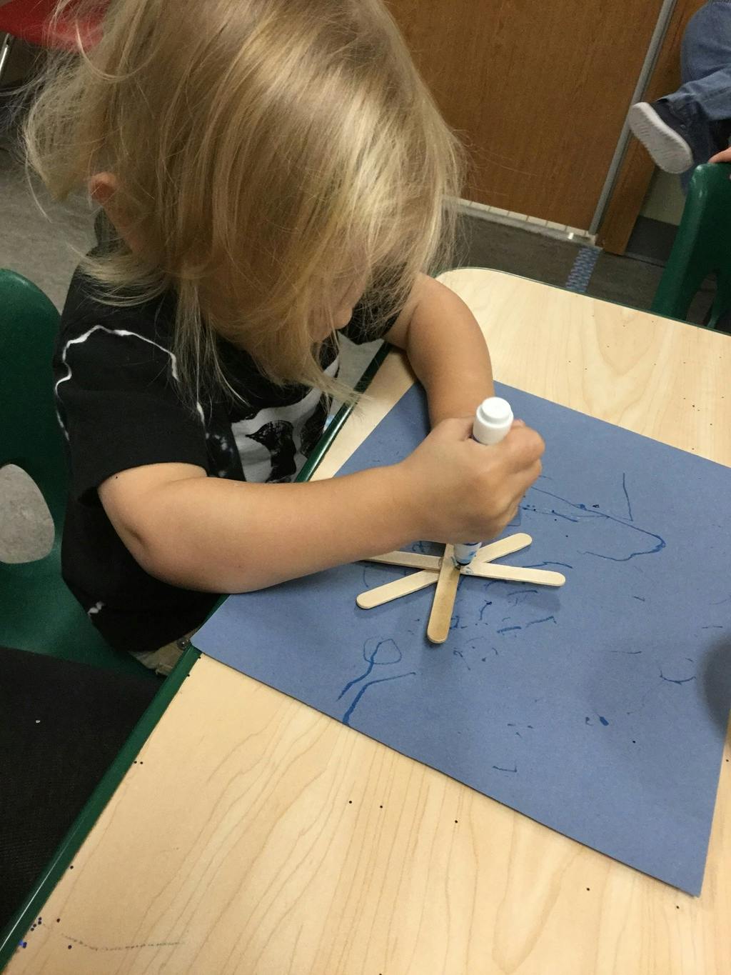 Child gluing craft sticks onto blue paper in a classroom