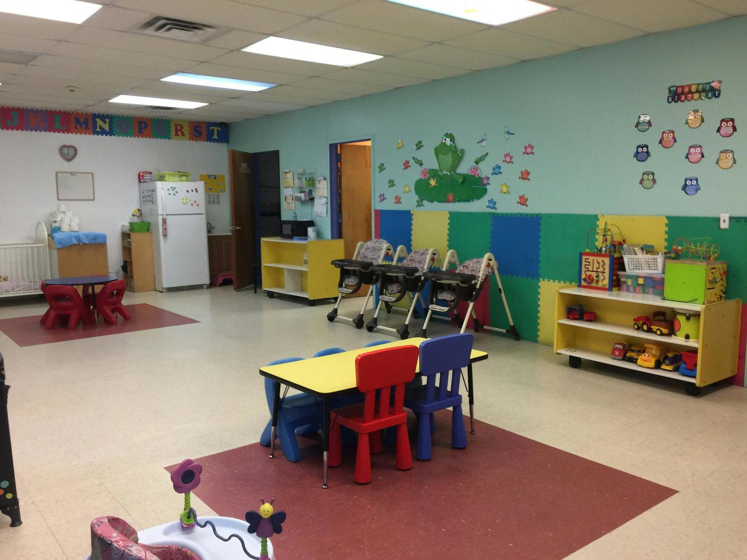 A colorful classroom with small tables and chairs, high chairs, and shelves with toys