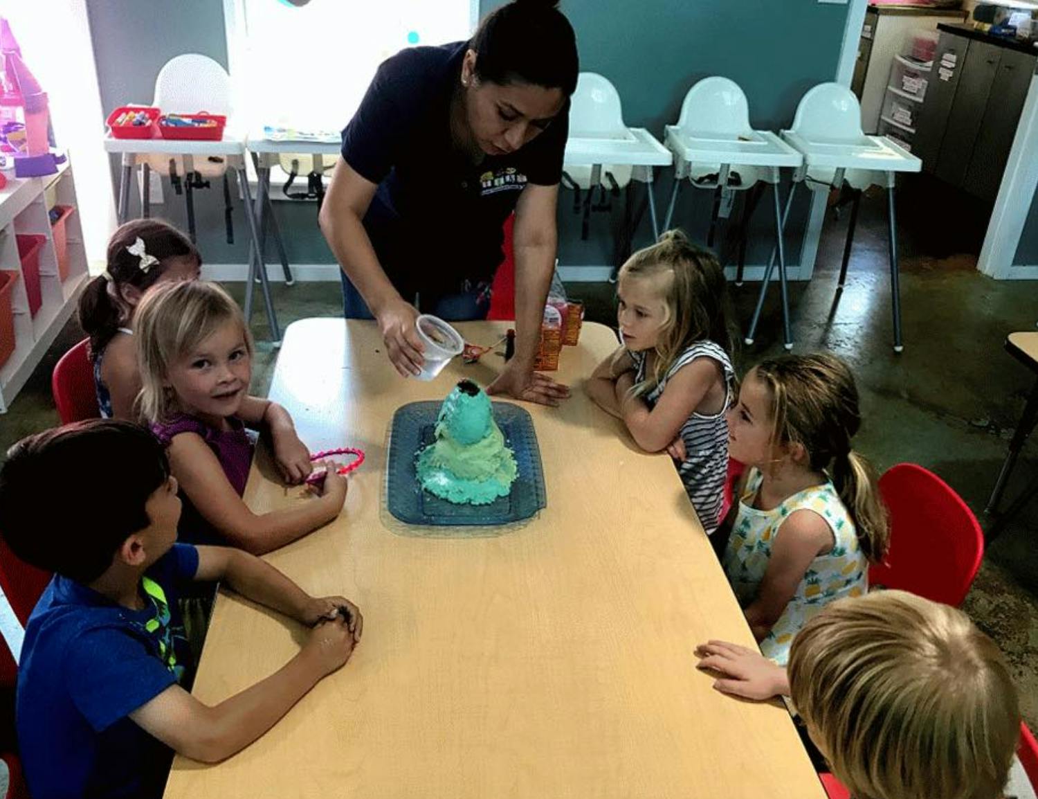 Person pouring liquid into a volcano model with children watching at a table