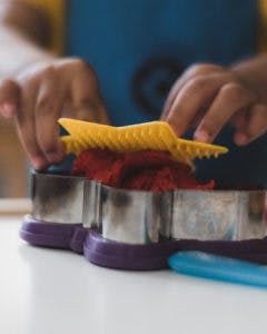 Child's hands pressing red dough with a yellow tool into a cookie cutter