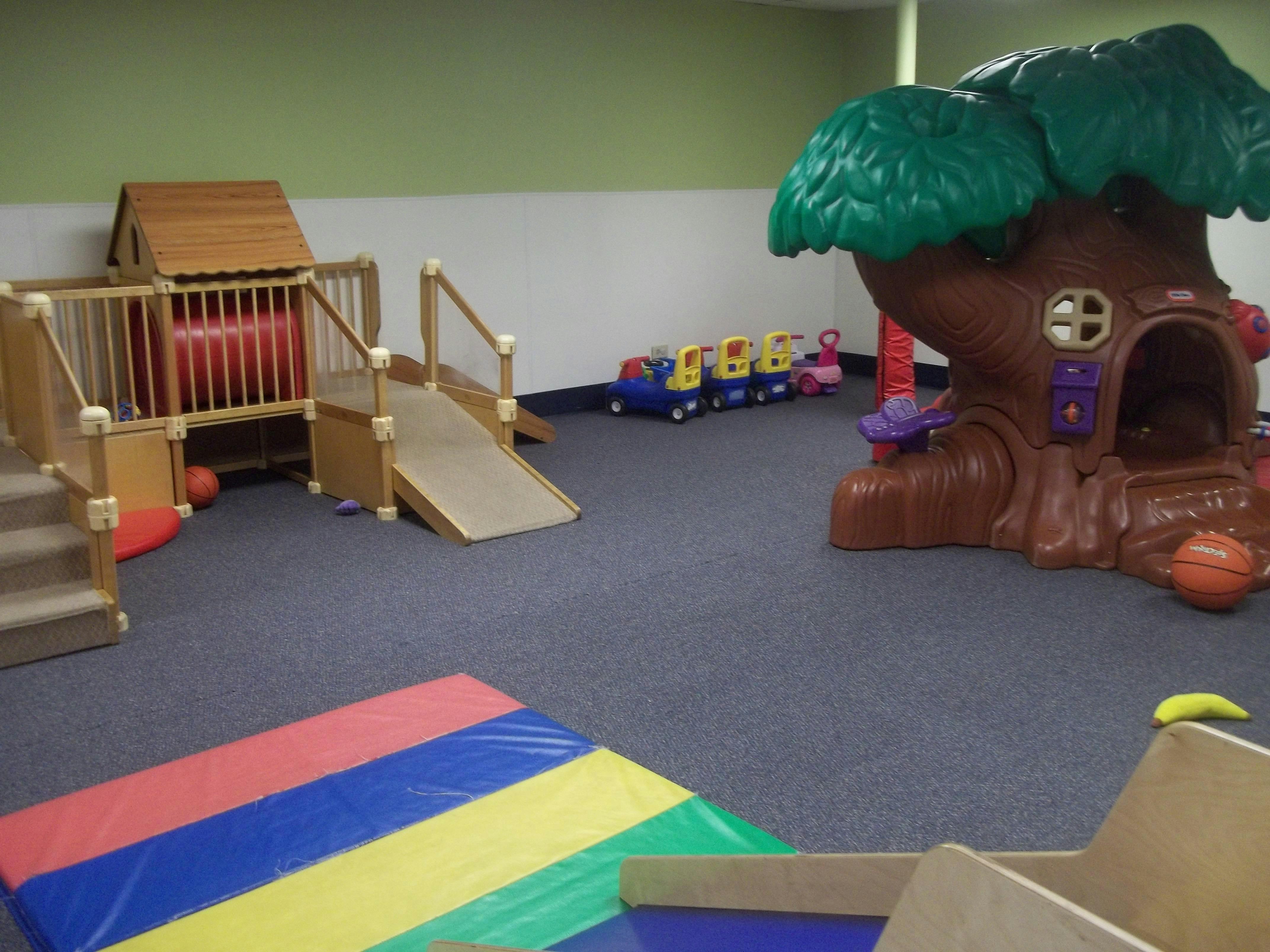 Indoor play area with a wooden play structure, a plastic tree playhouse, and ride-on toys