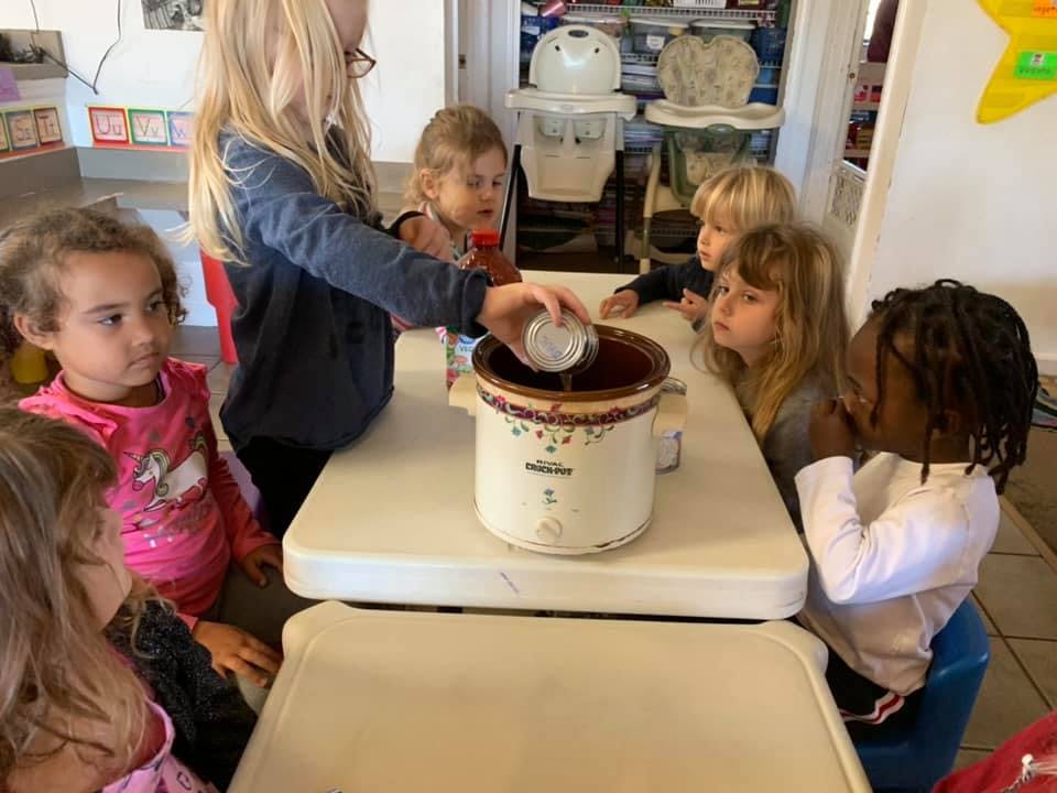 Children gathered around a table, one pouring from a cup into a slow cooker