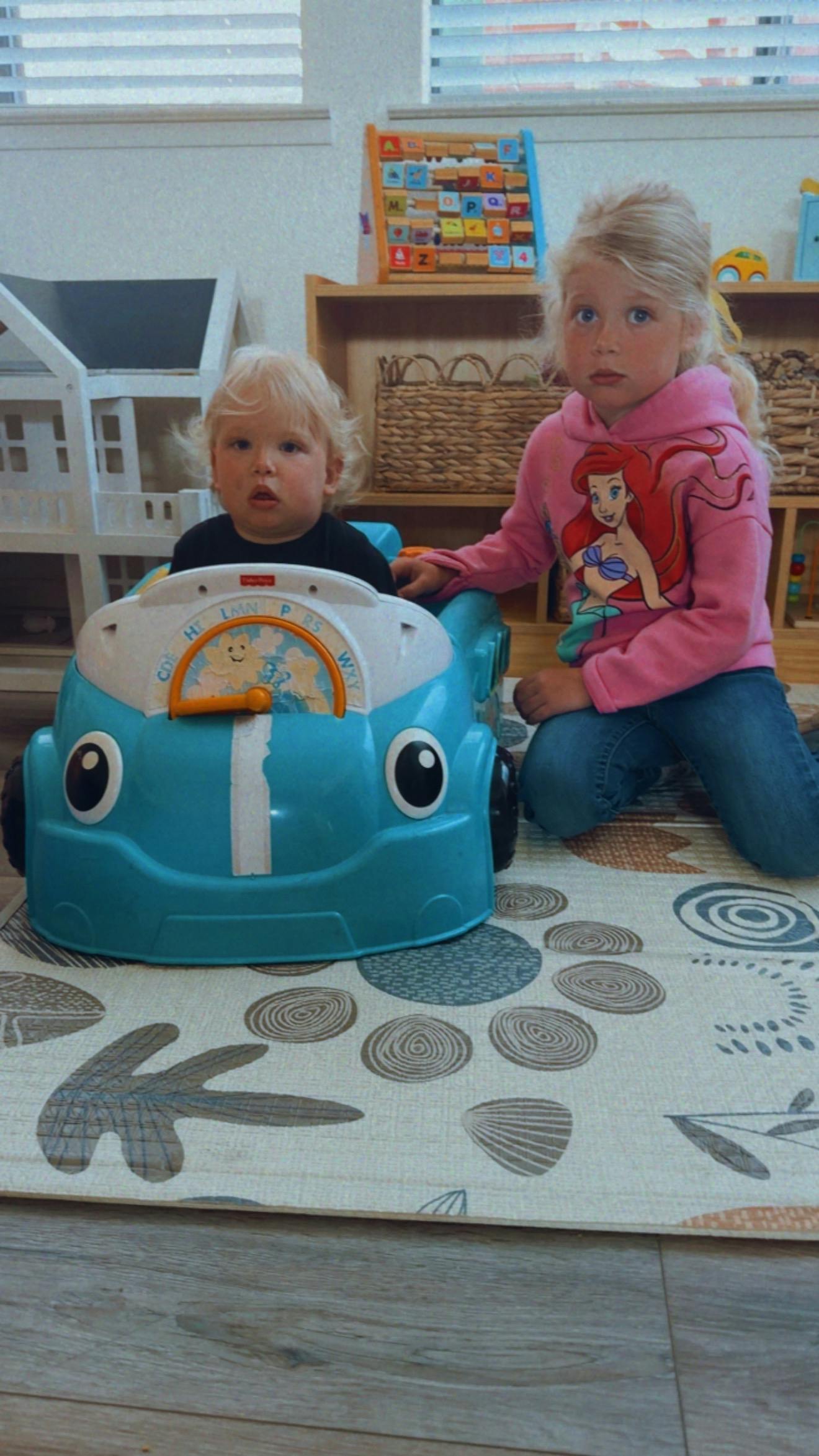 Child sitting in a blue toy car with another child kneeling beside it
