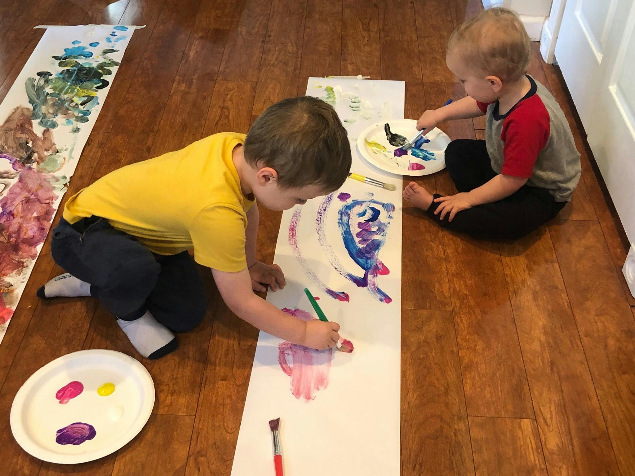 Two children painting on long sheets of paper on a wooden floor