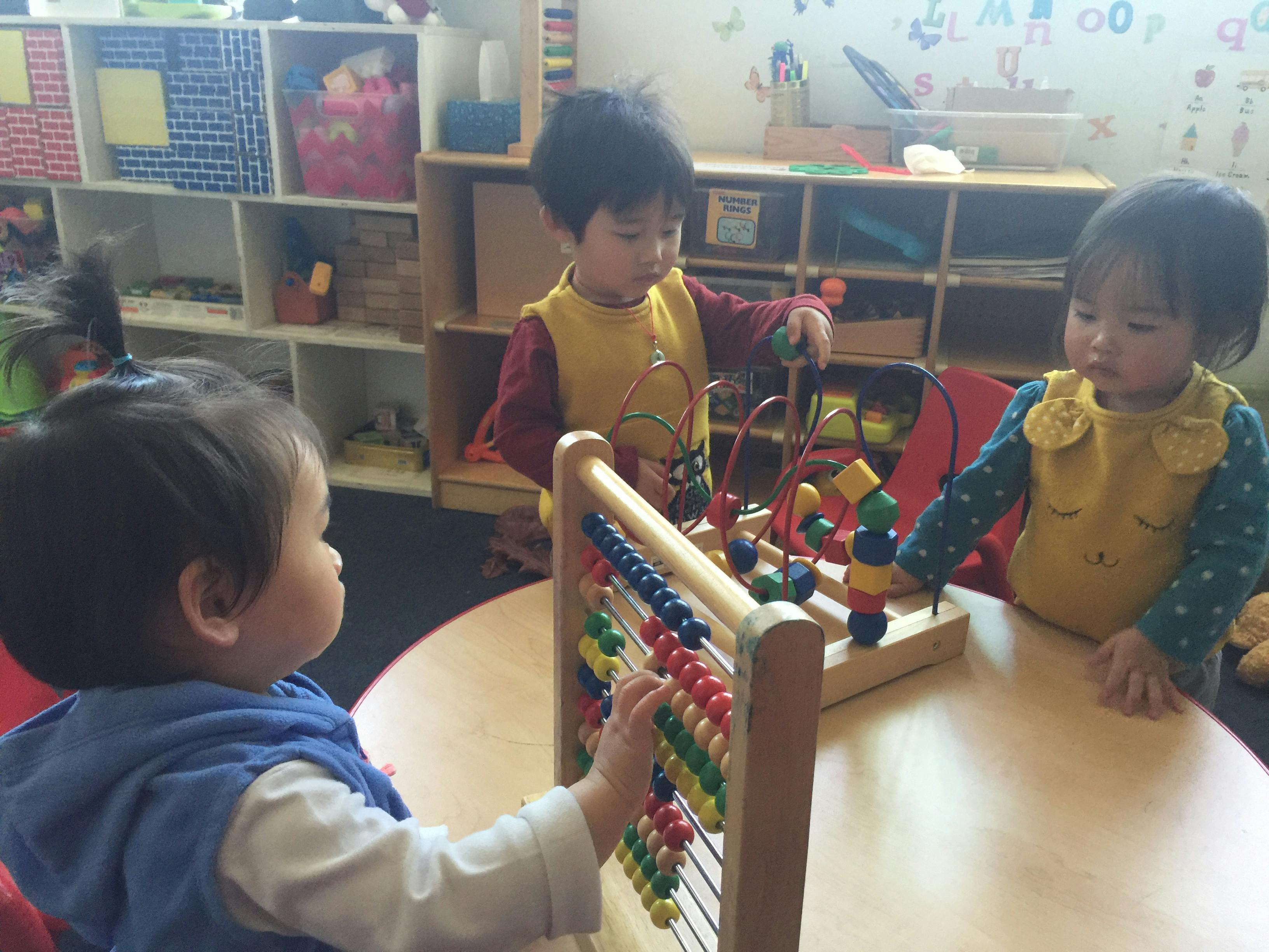 Three children playing with an abacus and a bead maze toy in a classroom