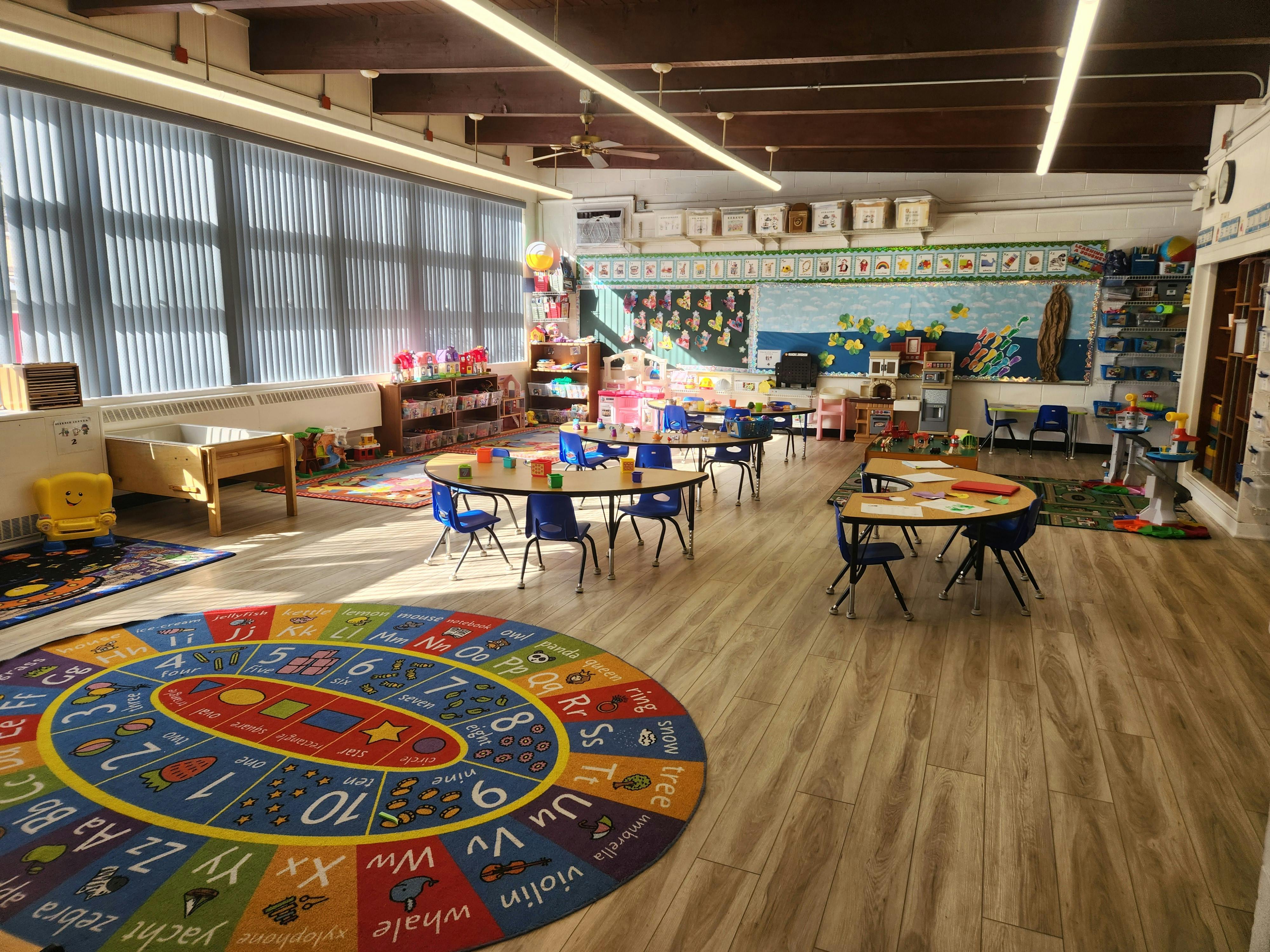 Classroom with small tables and chairs, a colorful alphabet rug, and large windows