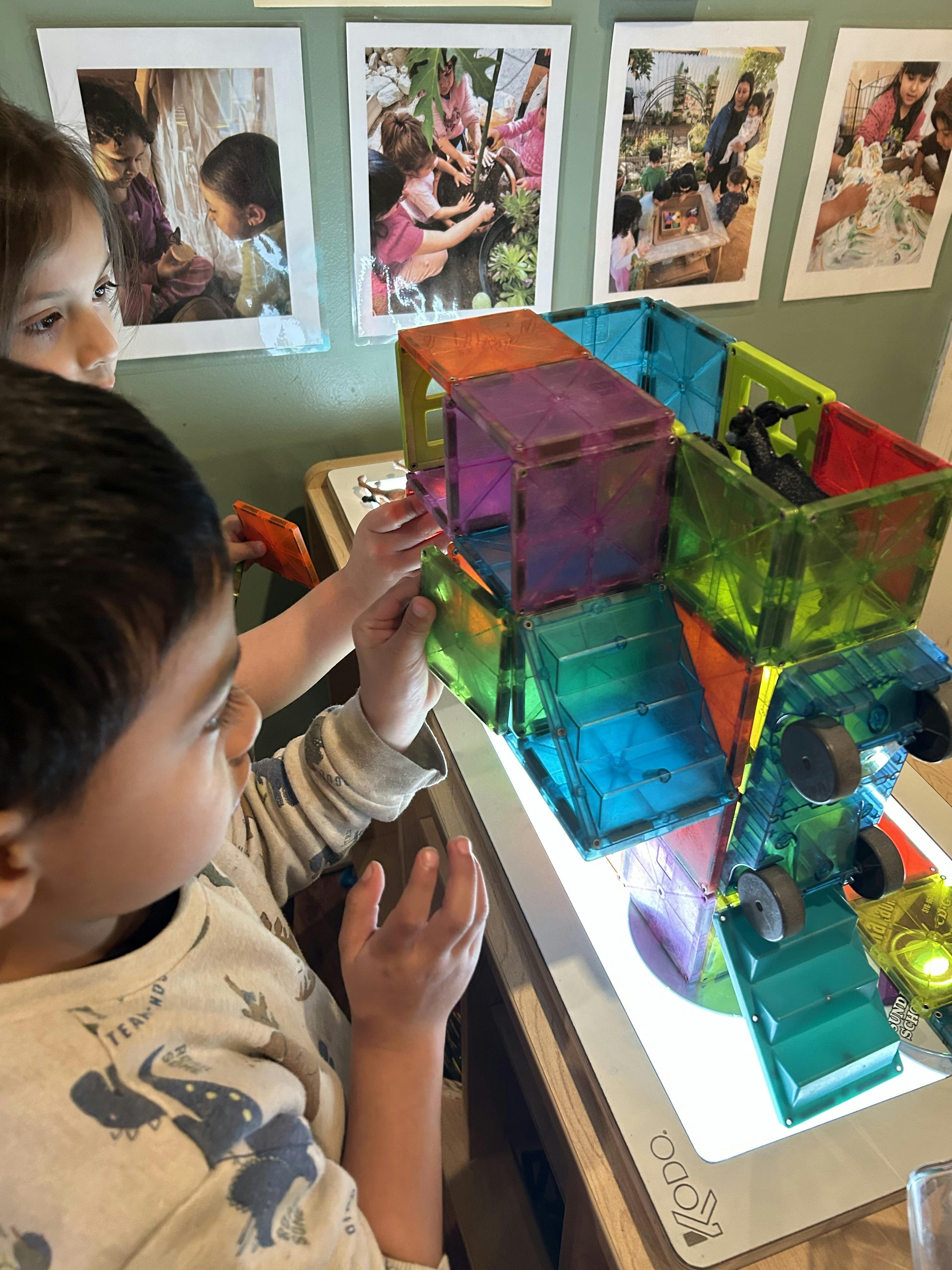 Two children building a structure with magnetic tiles on a light table