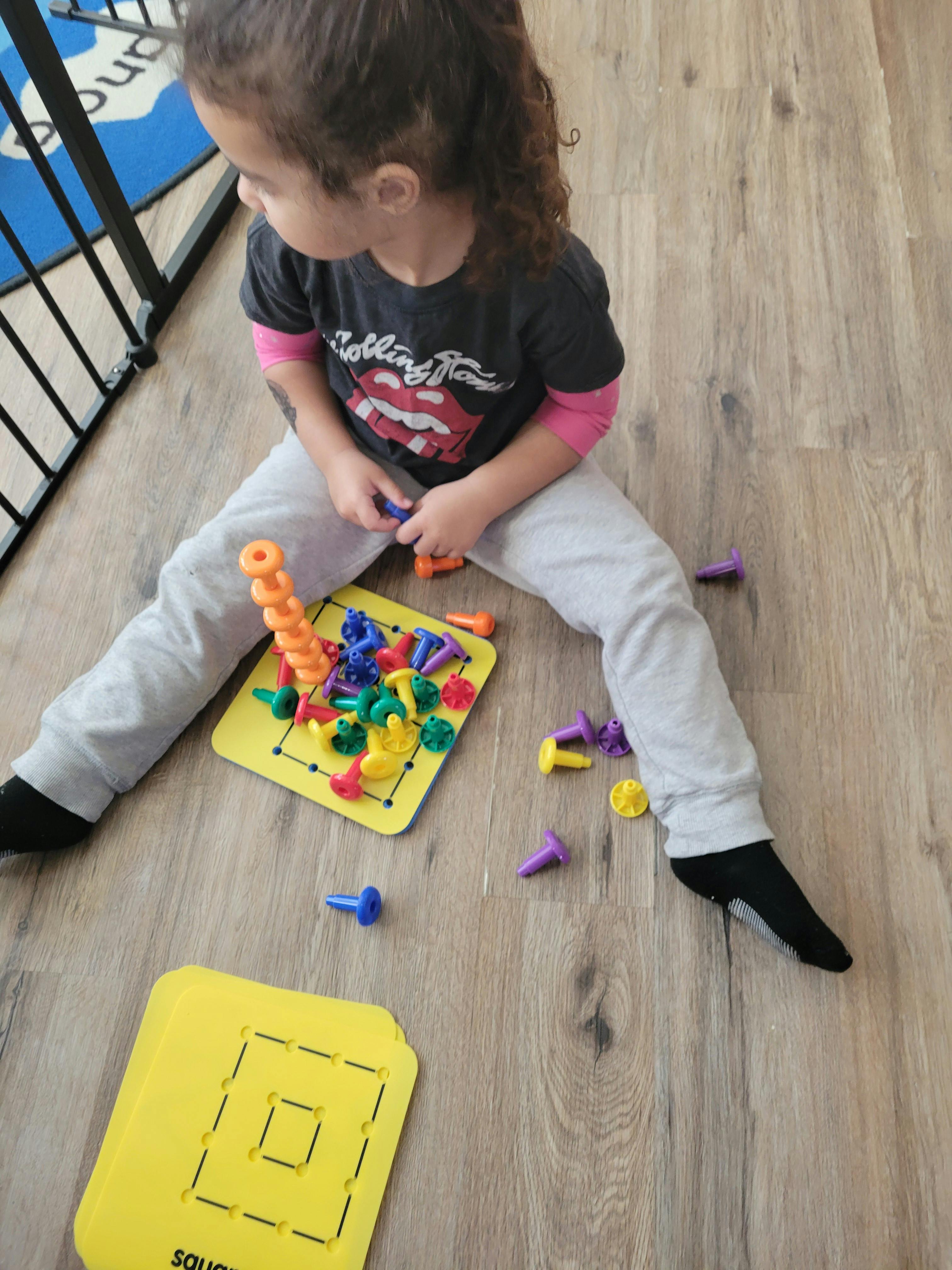 Child sitting on a wooden floor stacking colorful plastic pegs on a pegboard