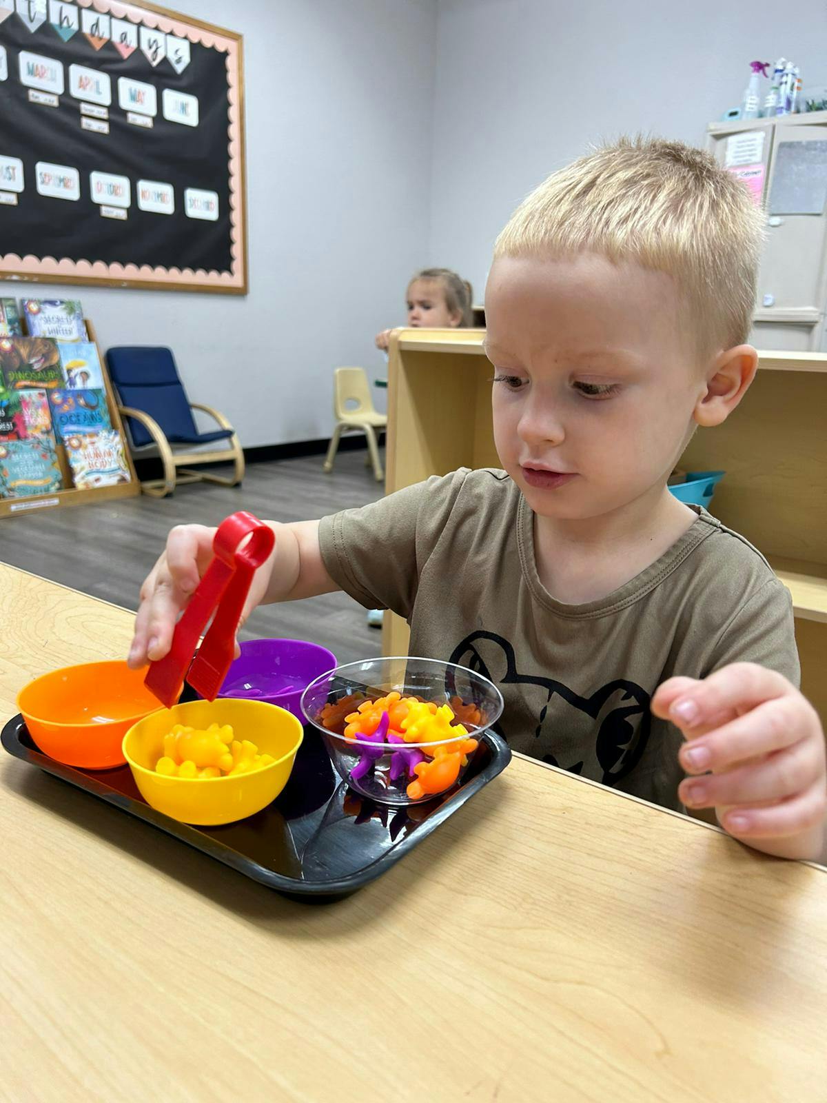 Child using tongs to move small objects between bowls in a classroom