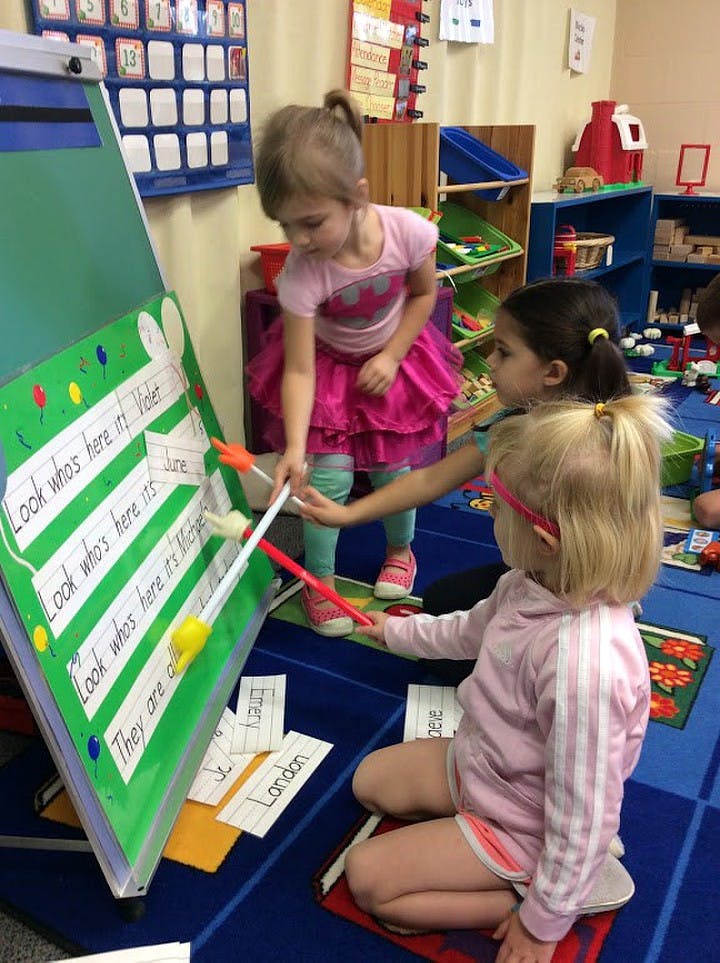 Three children using pointers on a green easel with text in a classroom