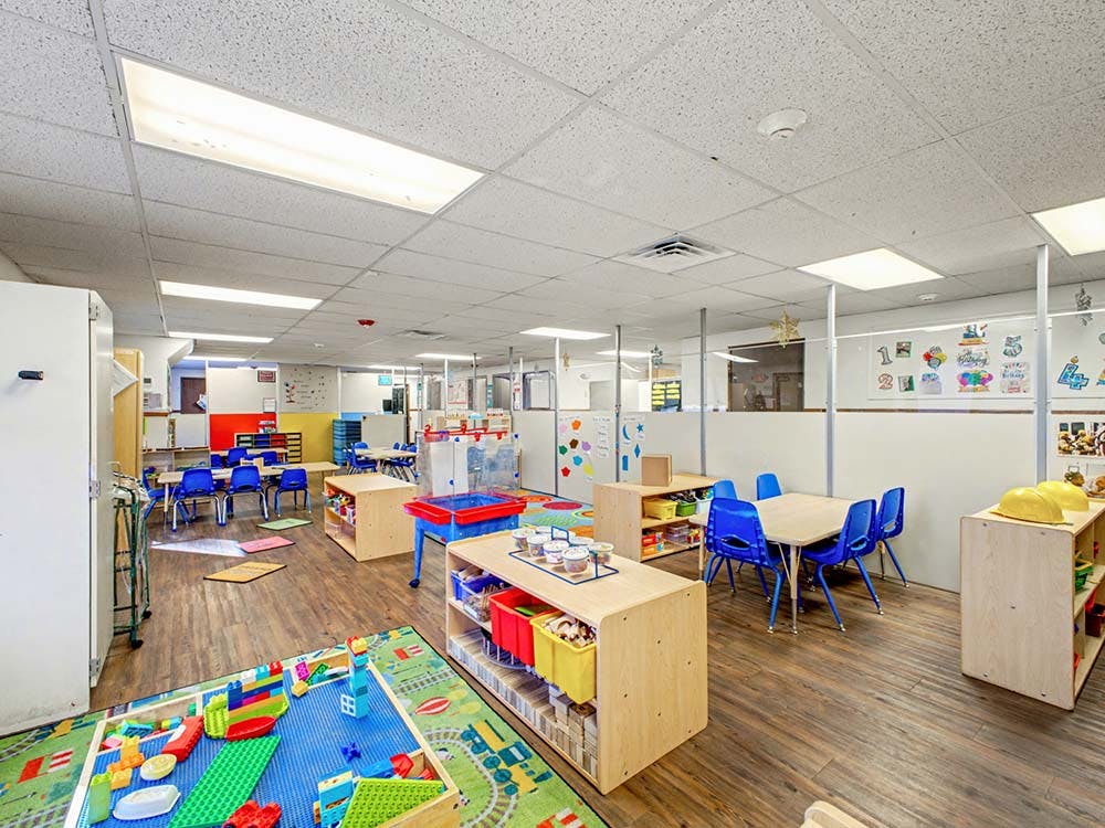 Classroom with tables, blue chairs, shelves, and a colorful rug with building blocks