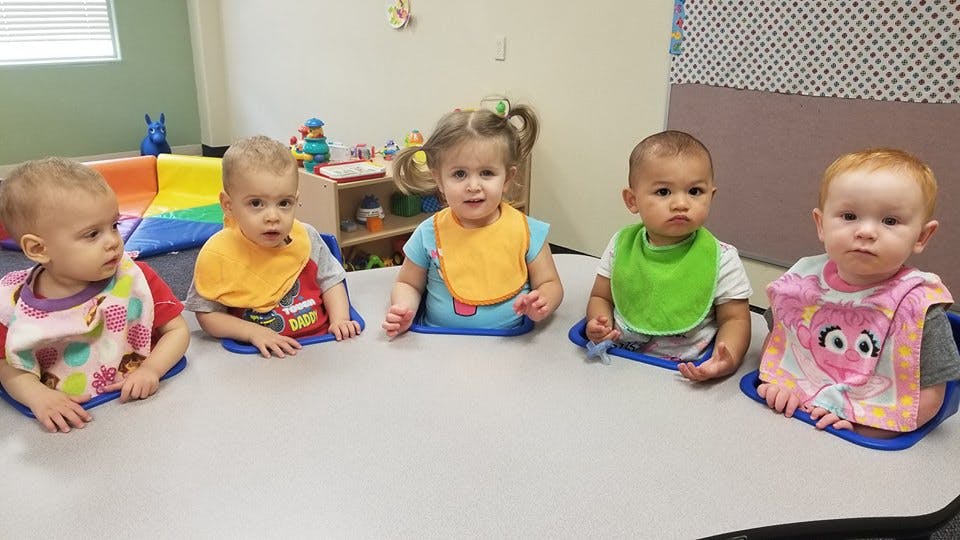 Five babies wearing bibs sitting at a table